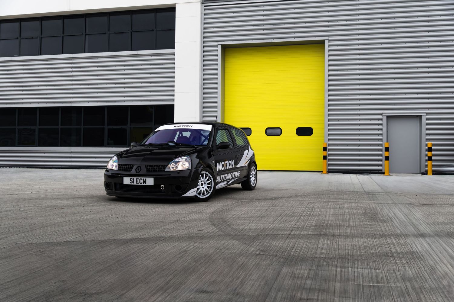 Black Renault Clio track car with Motion decals parked on concrete outside yellow industrial doors.