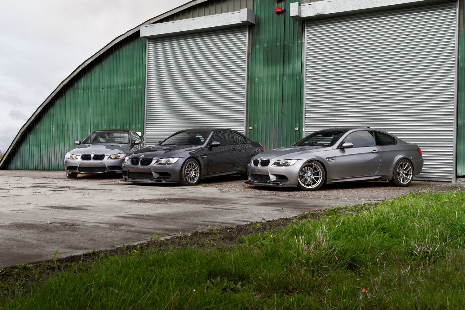 Three grey BMW M3 coupes lined up outside a curved green industrial building.