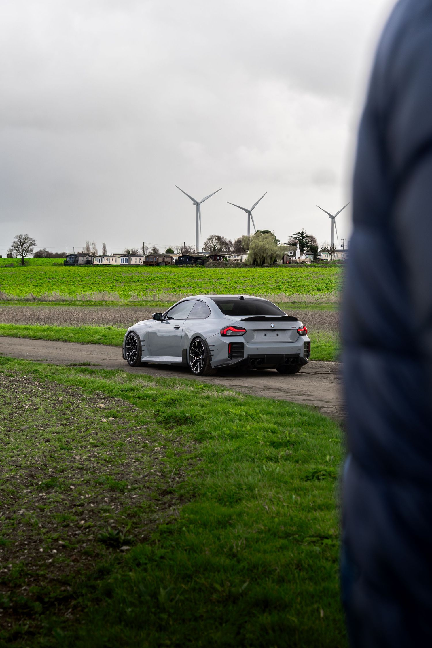 Silver BMW M2 coupe photographed from behind on a rural road with wind turbines in the green landscape.