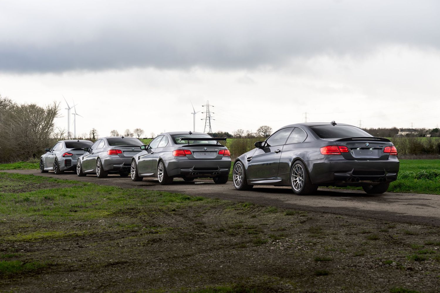 Five BMW M3 coupes lined up on a grassy rural area under a moody overcast sky.