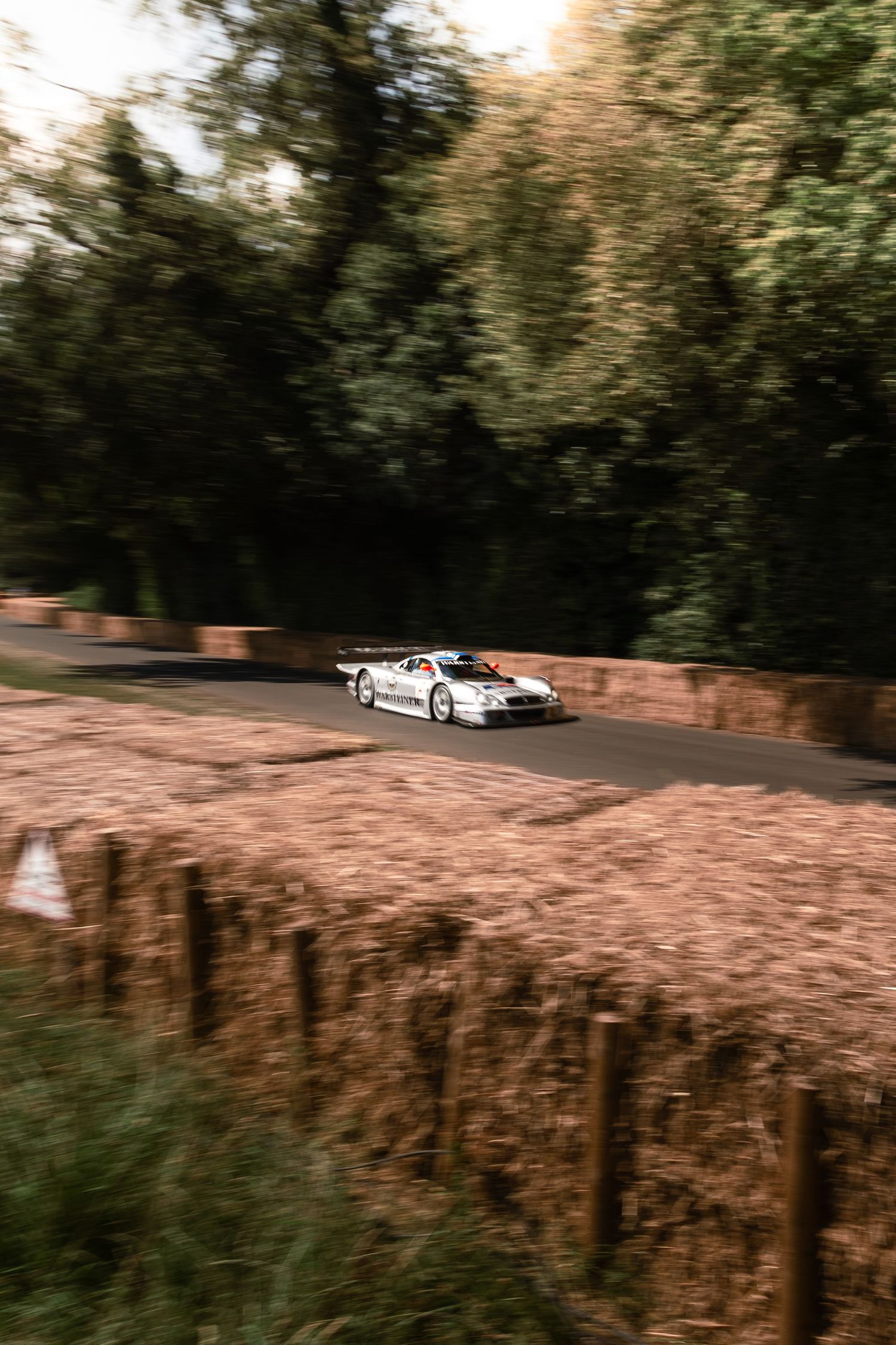 White racing car speeding past hay bale barriers on a tree-lined hillclimb course.