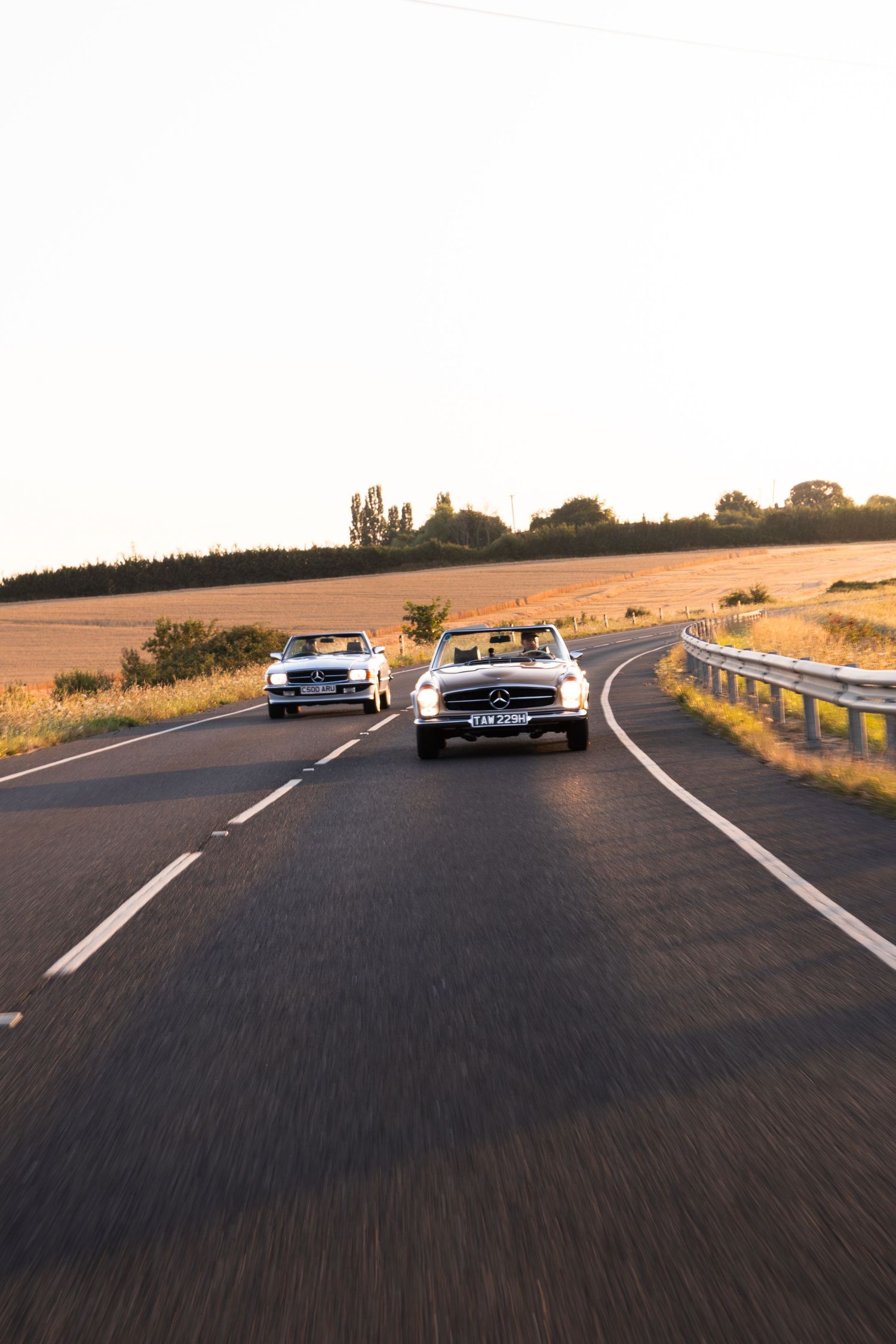 Two classic Mercedes-Benz convertibles driving on a rural road at golden hour sunset.