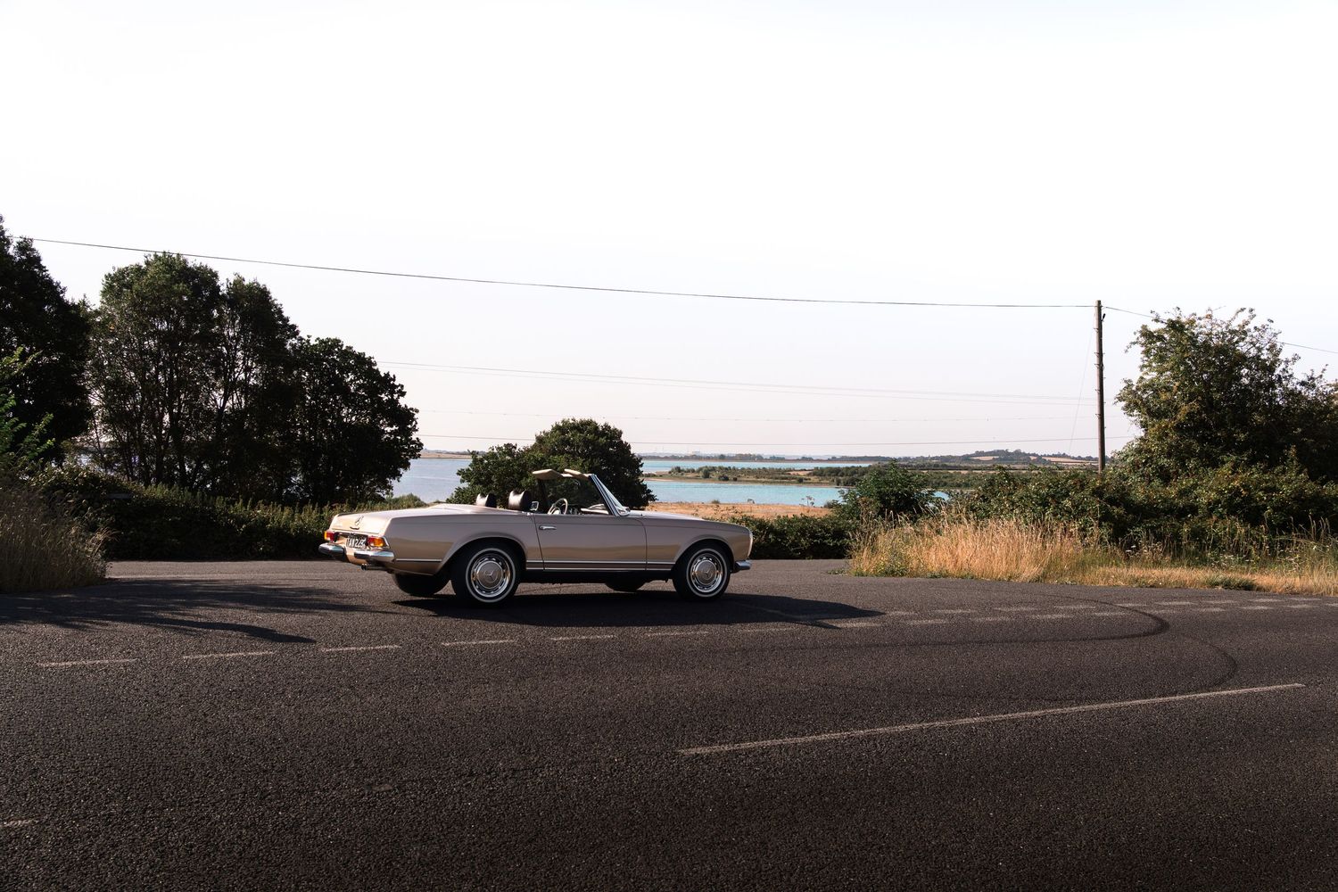 Vintage white convertible car parked on a quiet road surrounded by trees and water views on a calm day.