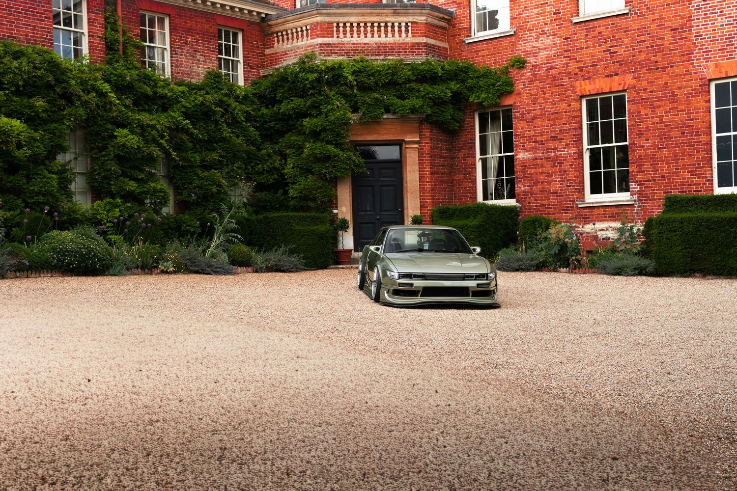 A sleek green sports car parked on a gravel driveway in front of a grand red brick manor house with ivy.