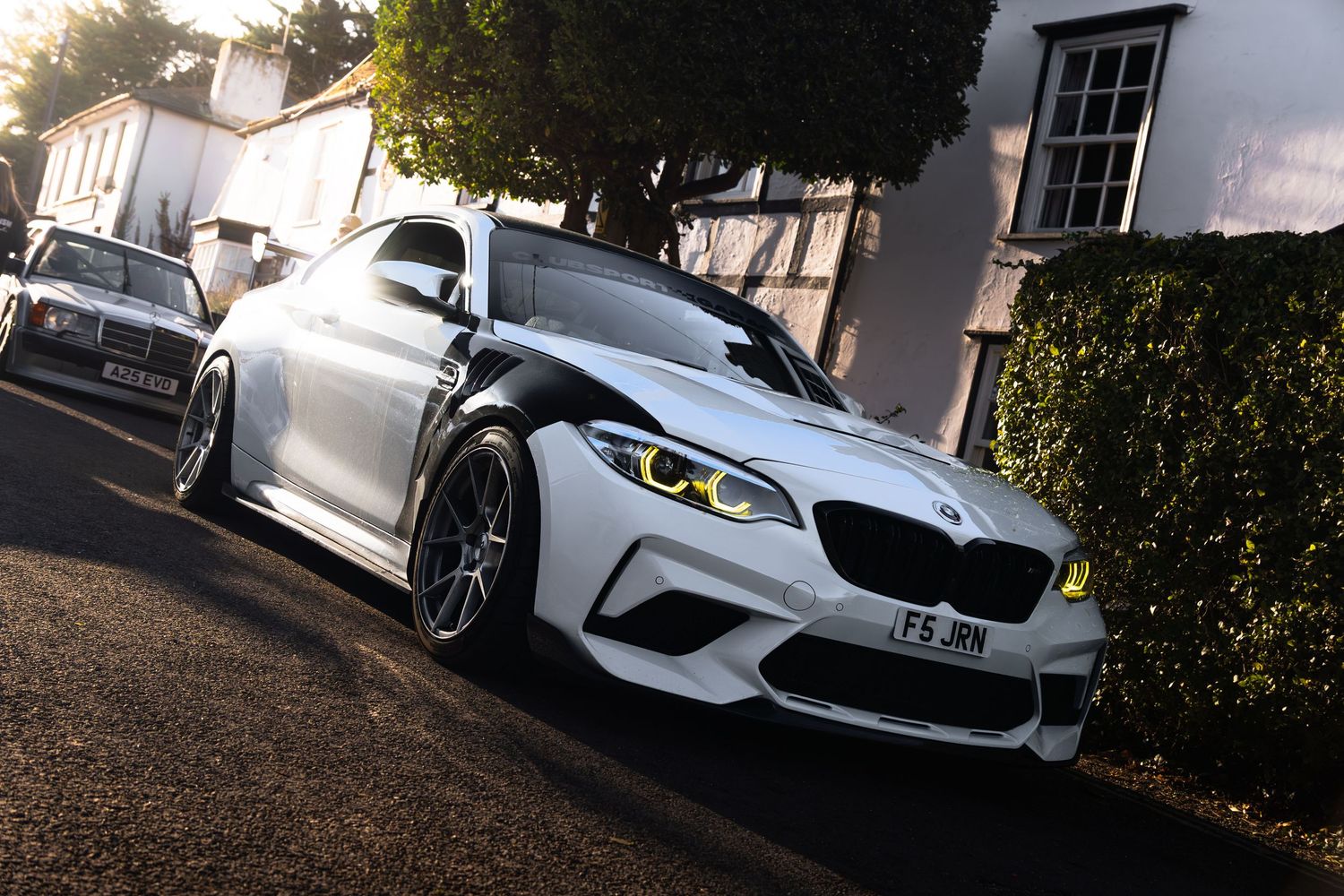 White BMW M2 with yellow angel eyes and wide body kit parked on a residential street.