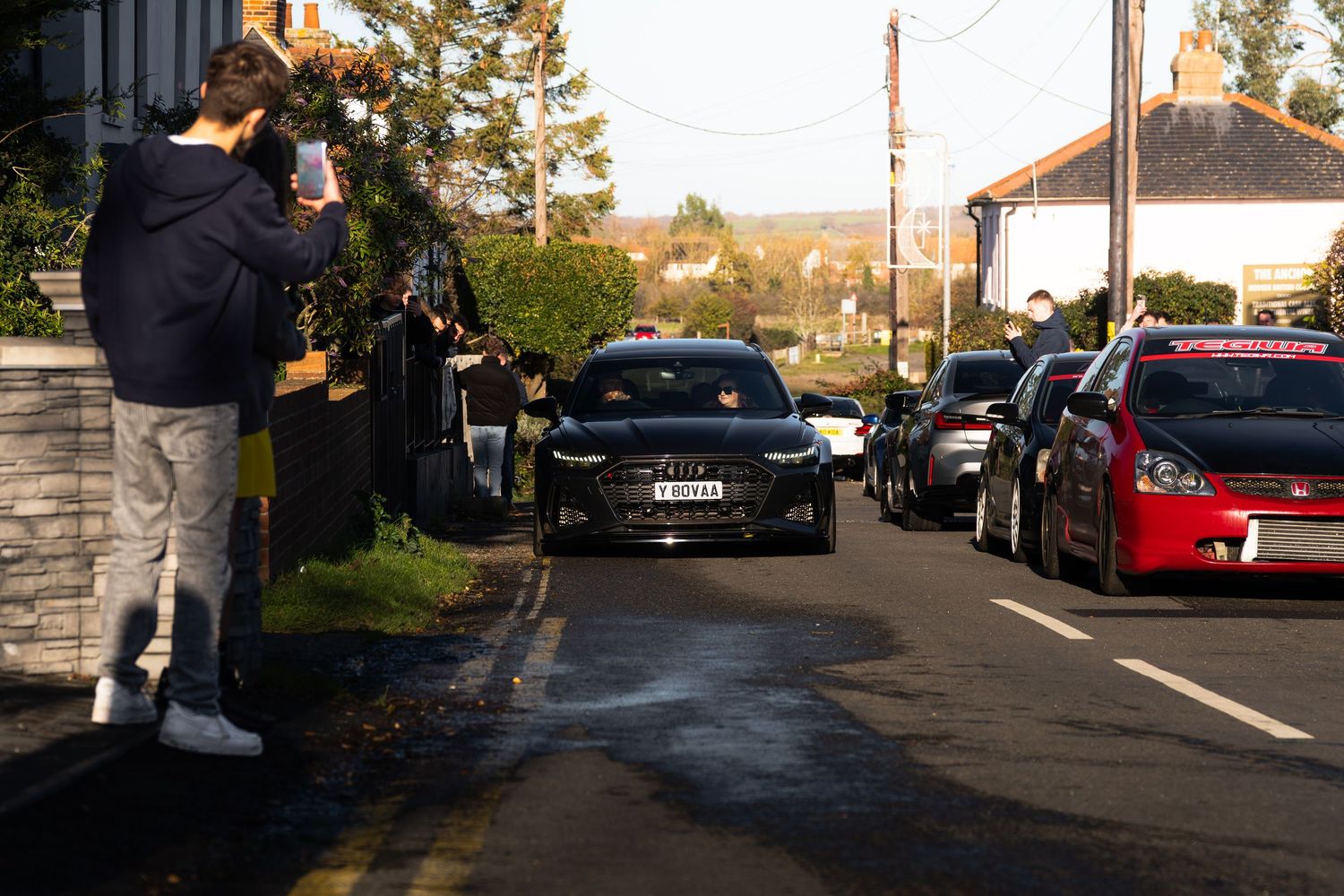 A black sedan driving down a residential street while a photographer captures the moment.