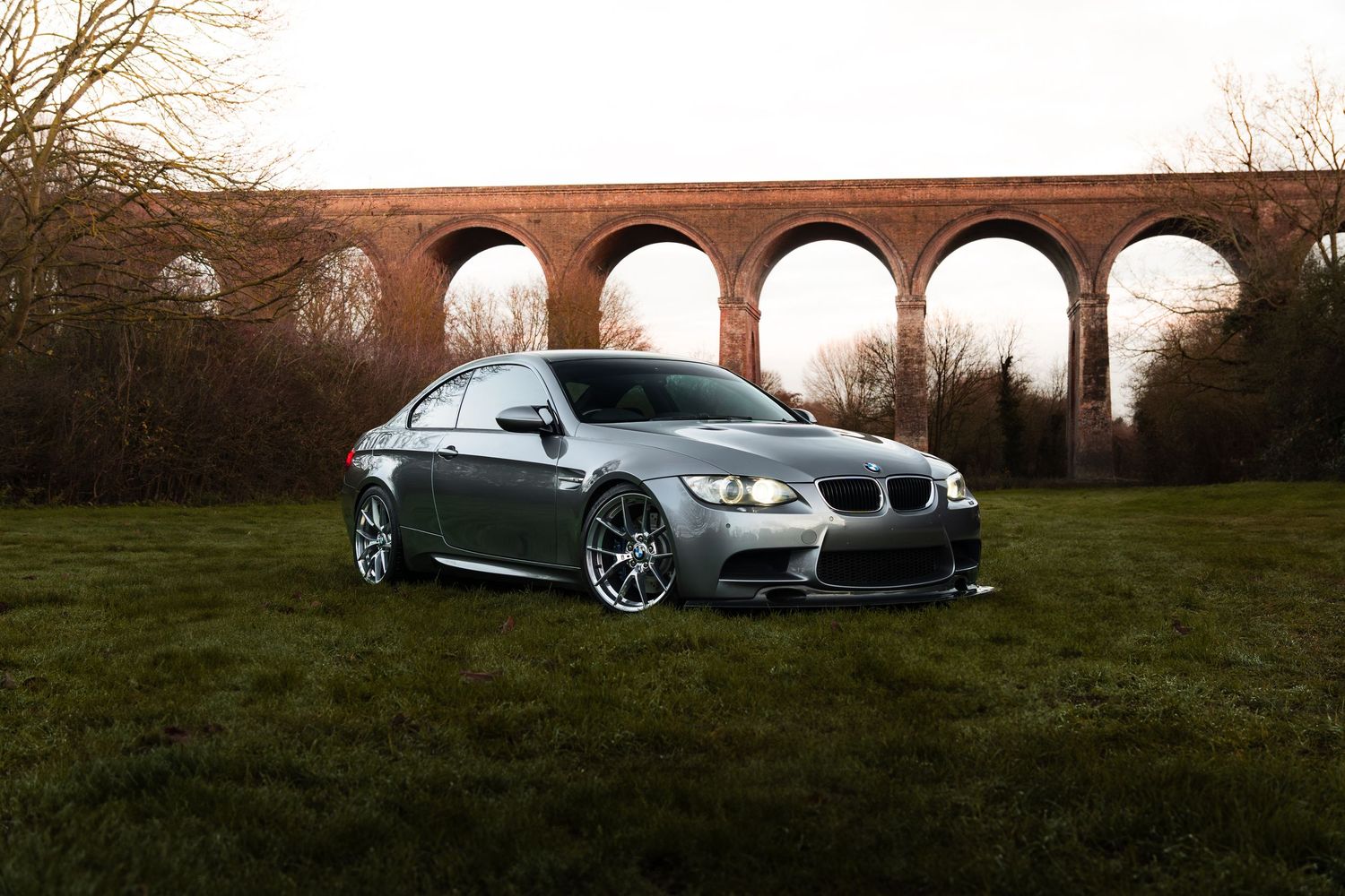 A silver BMW M3 coupe with aftermarket wheels posed on grass before a brick viaduct.