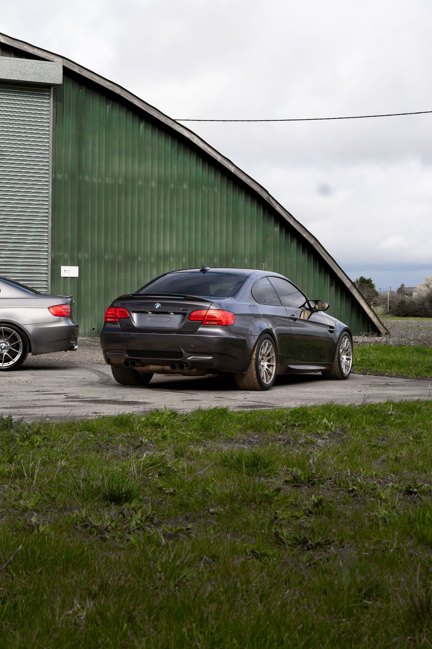 Dark gray BMW M3 coupe parked from rear view beside a green corrugated metal hangar.