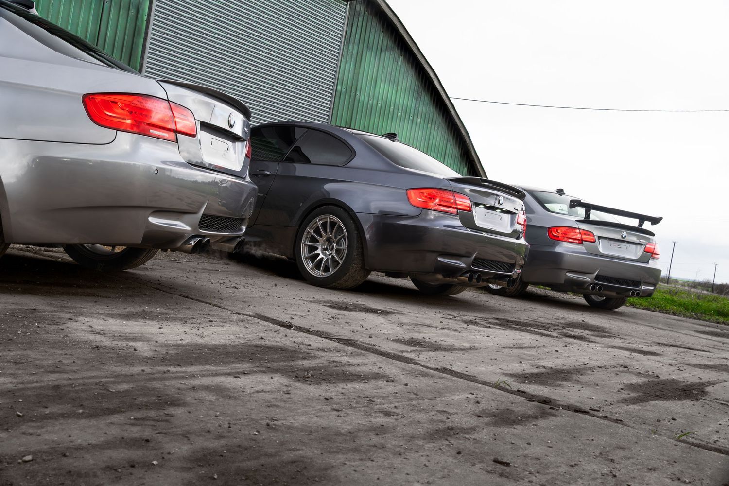 Three BMW M3 cars parked at an angle beside a green corrugated metal hangar building.