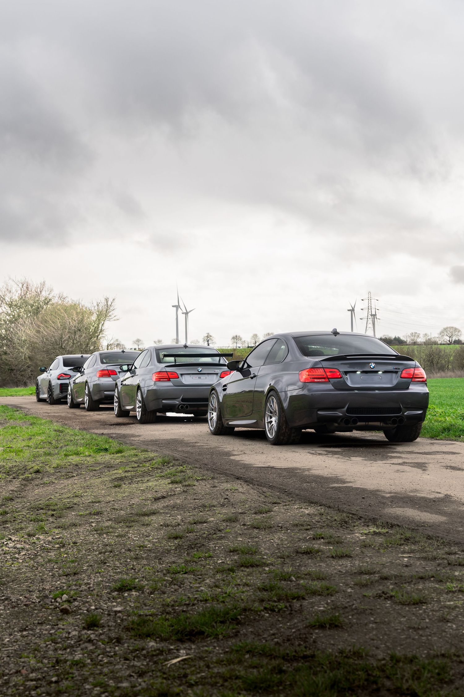 Row of dark BMW M3 coupes lined up on a rural dirt road with wind turbines visible in the cloudy background.