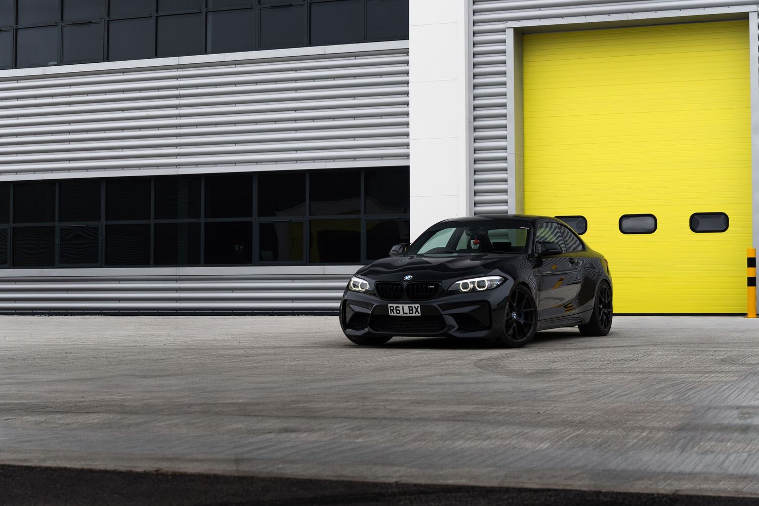 Black BMW M2 coupe parked at an angle in front of large yellow garage doors.