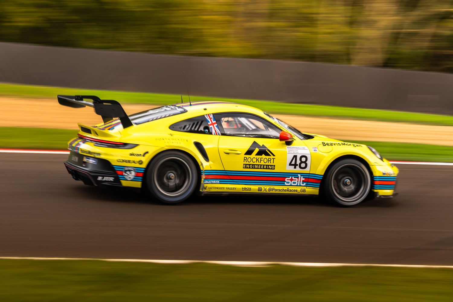 Yellow Porsche 911 GT3 number 48 with retro livery racing on track with autumn trees backdrop.