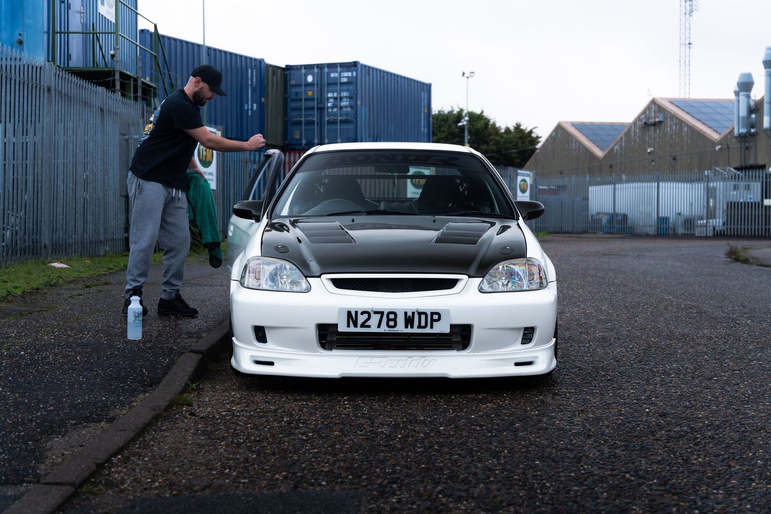A modified Honda Civic with white bumper kit and carbon hood being cleaned in a parking lot.