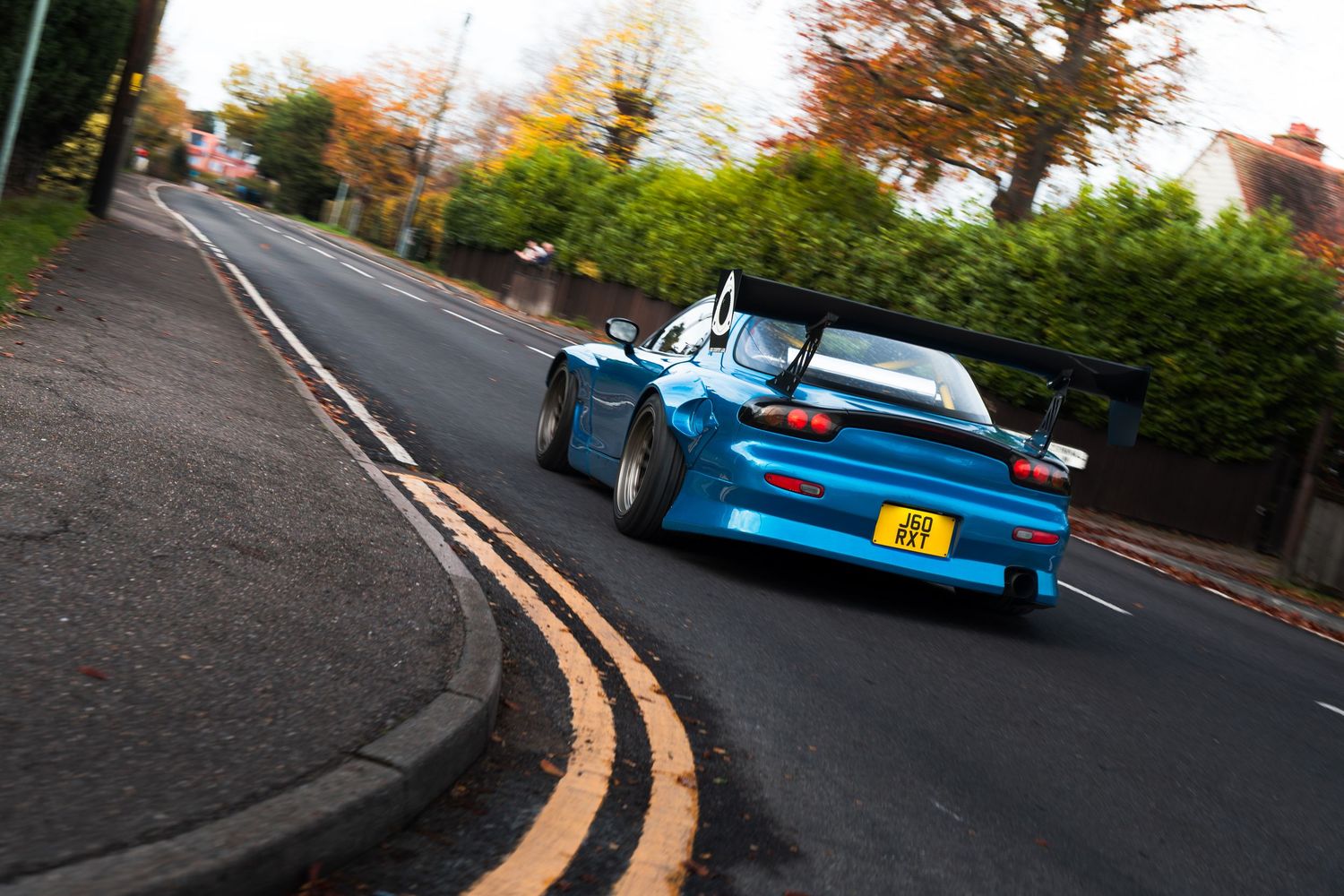 Blue widebody Mazda RX-7 with yellow number plate cornering on a tree-lined UK road.