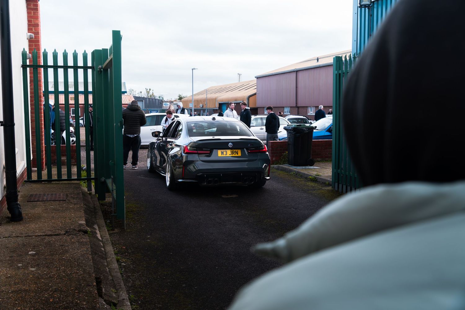Cars entering through a gate at an outdoor automotive meet with spectators watching nearby.