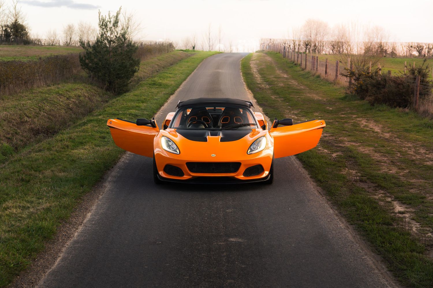 Orange Lotus Elise with butterfly doors open facing forward on a straight country road.