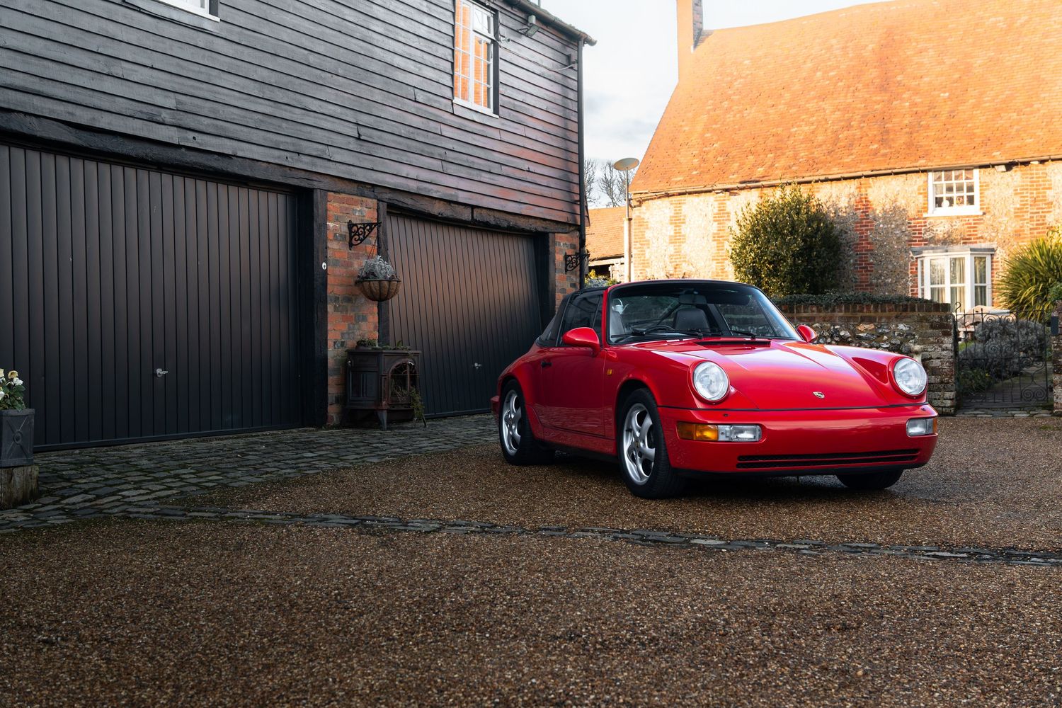 Red Porsche 911 Carrera 2 Targa parked on a sunlit gravel driveway beside a rustic stone farmhouse.