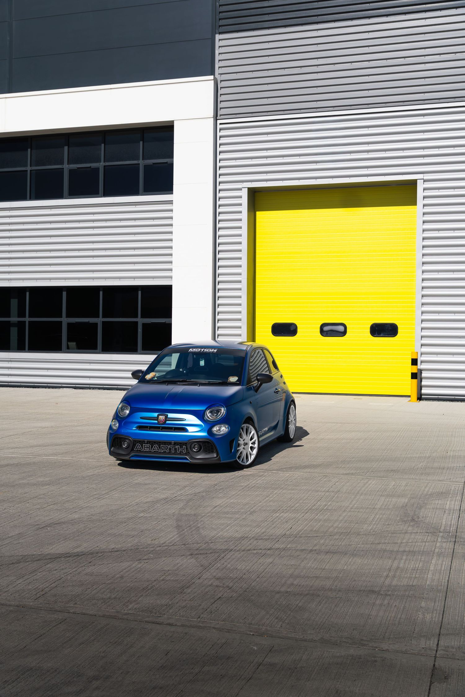 Blue Abarth 500 hot hatch photographed outside yellow industrial garage doors.