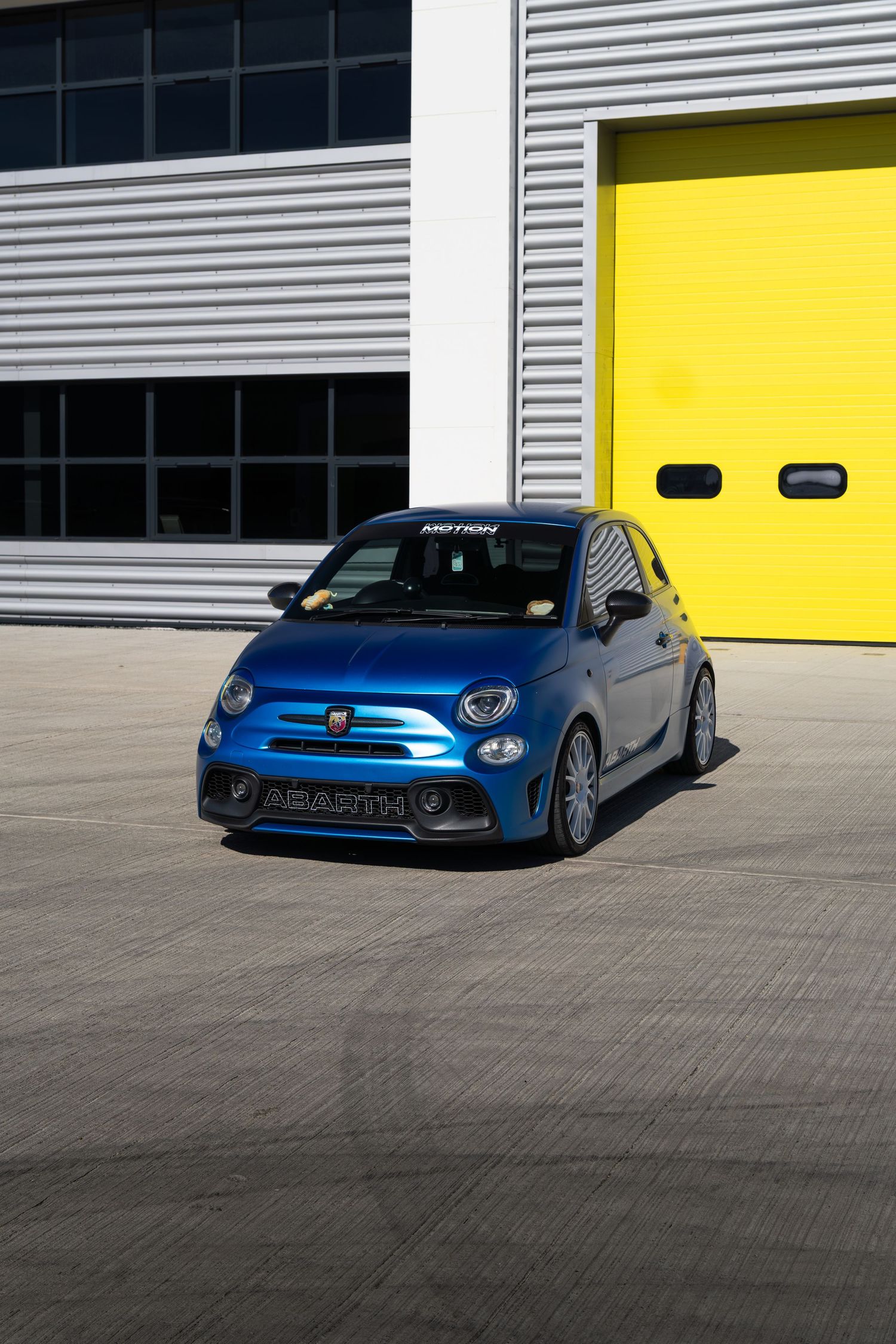 Blue Abarth 595 with Motion branding parked outside yellow industrial garage door.