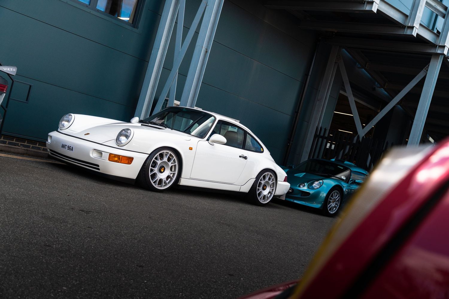 Classic white Porsche 911 parked outside a grey industrial building with a teal sports car nearby.