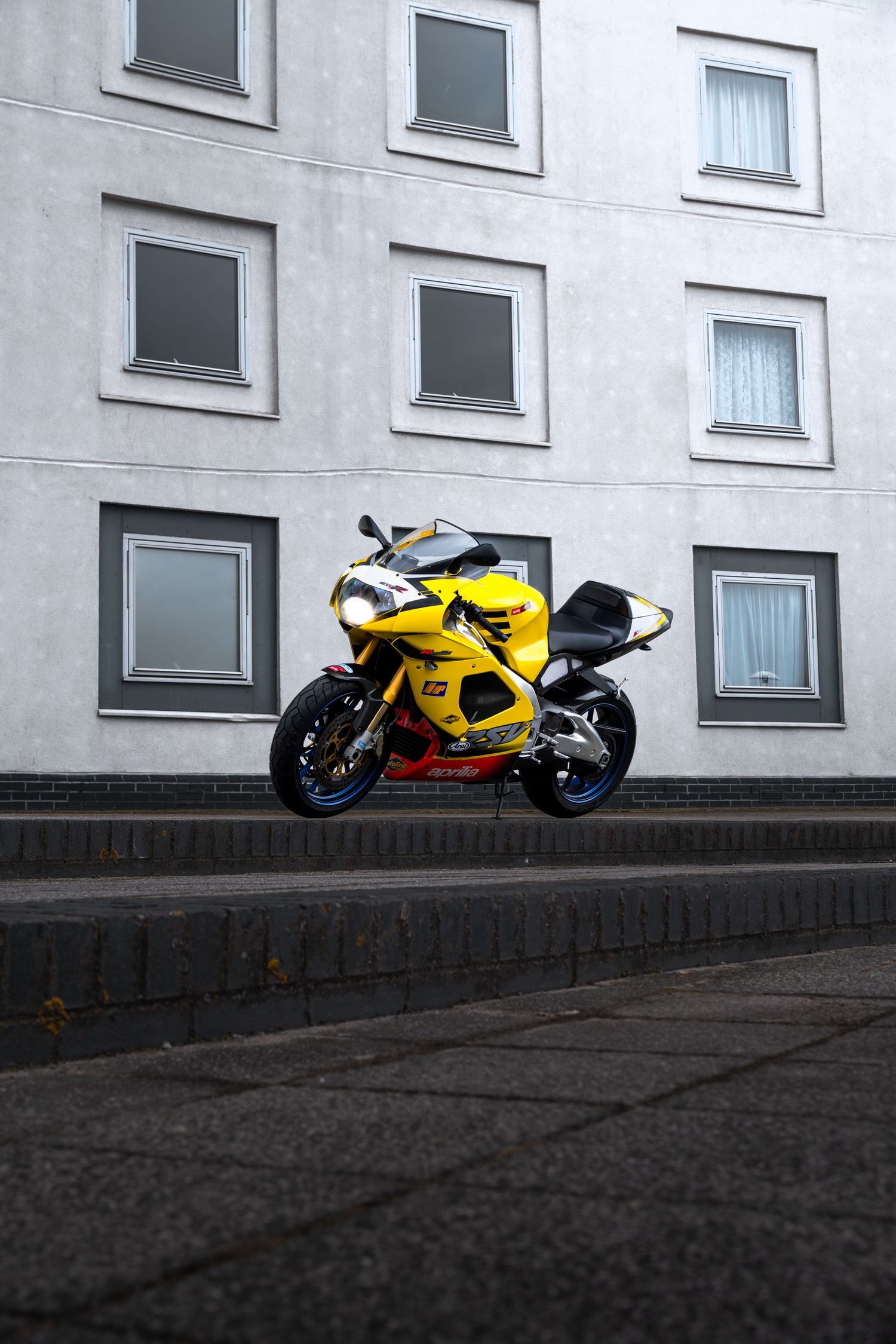 Yellow Aprilia RSV1000 sportbike parked on cobblestone steps in front of a gray residential building.