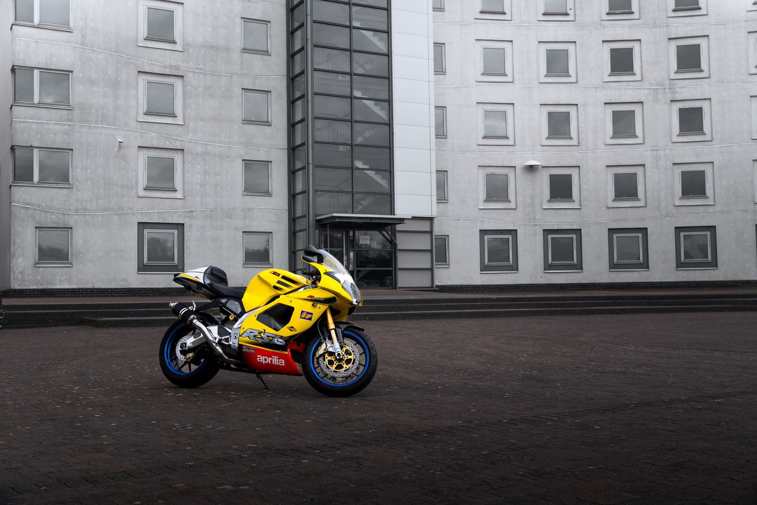 Yellow Aprilia RSV-R sport motorcycle displayed against a grey multi-windowed building exterior.