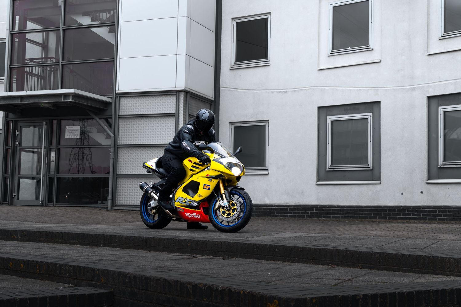 Rider on yellow Aprilia RSV superbike cruising past modern white building.