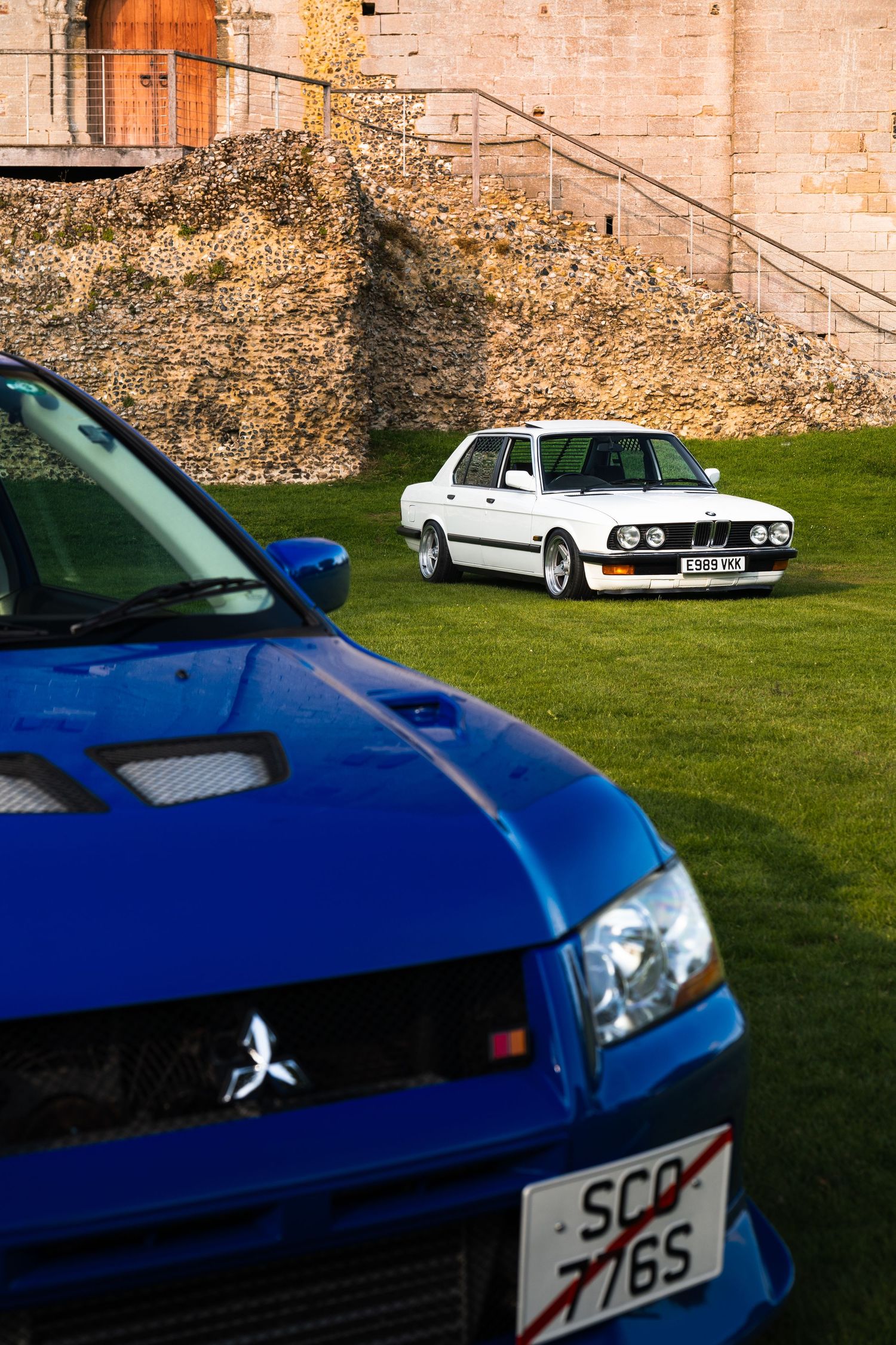 Blue Mitsubishi Evo and white BMW E28 parked on grass beside historic stone wall in Scotland.
