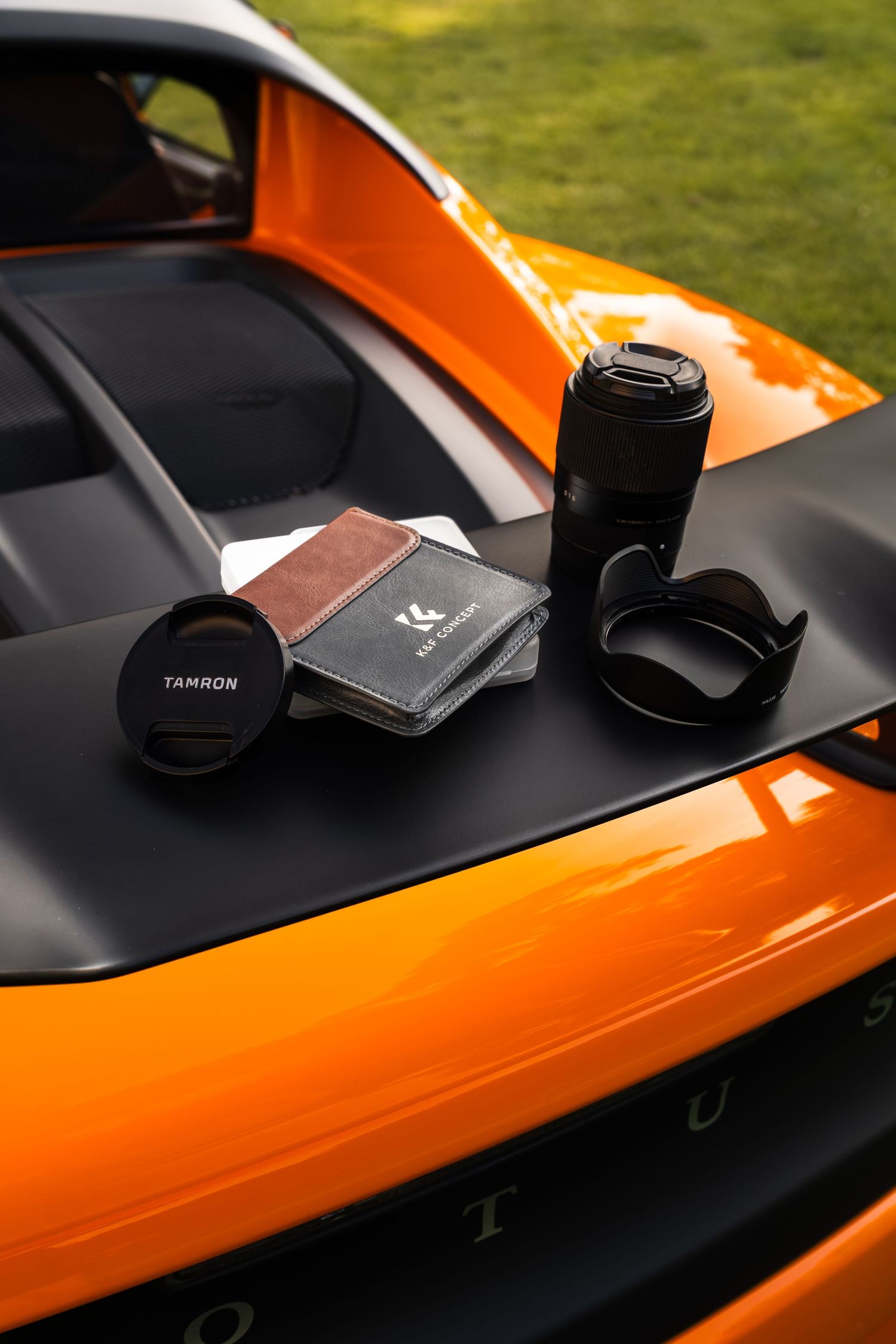 Tamron camera lens and accessories displayed on the hood of a vibrant orange Lotus sports car outdoors.