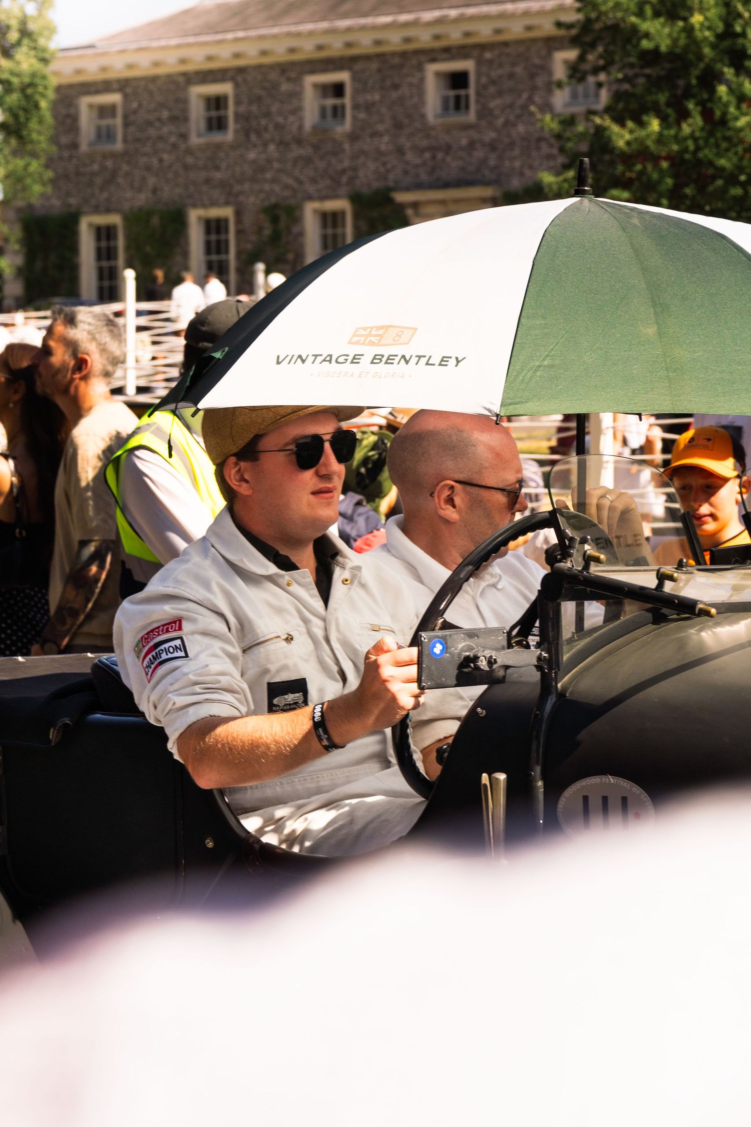 Two men in a vintage Bentley car at a classic car show, one holding a Vintage Bentley branded umbrella.
