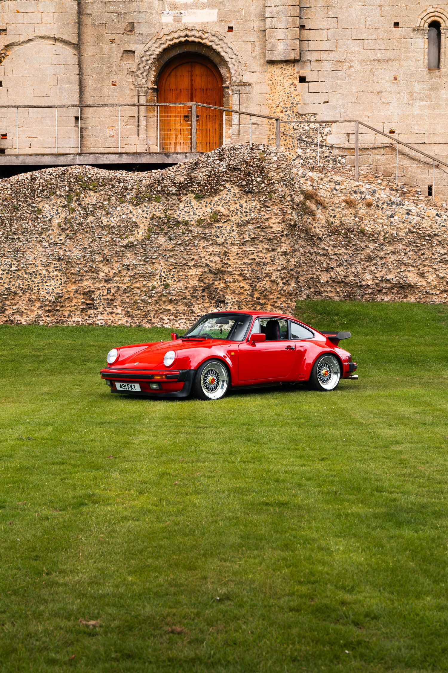 Red Porsche 911 Turbo parked on green lawn in front of historic stone castle with wooden arched door.