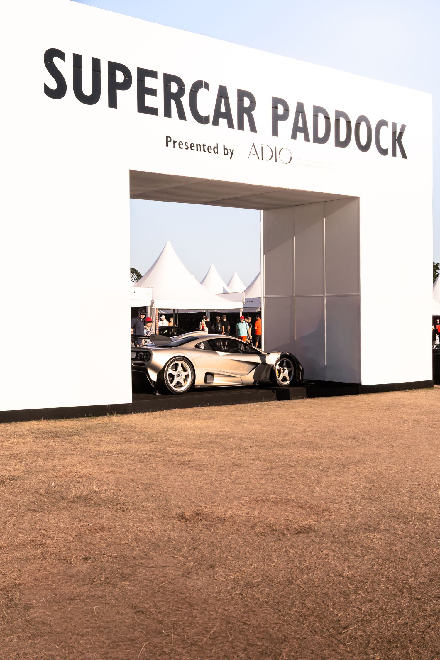 A silver supercar parked inside the Supercar Paddock entrance, presented by ADIO, at an outdoor motorsport event.