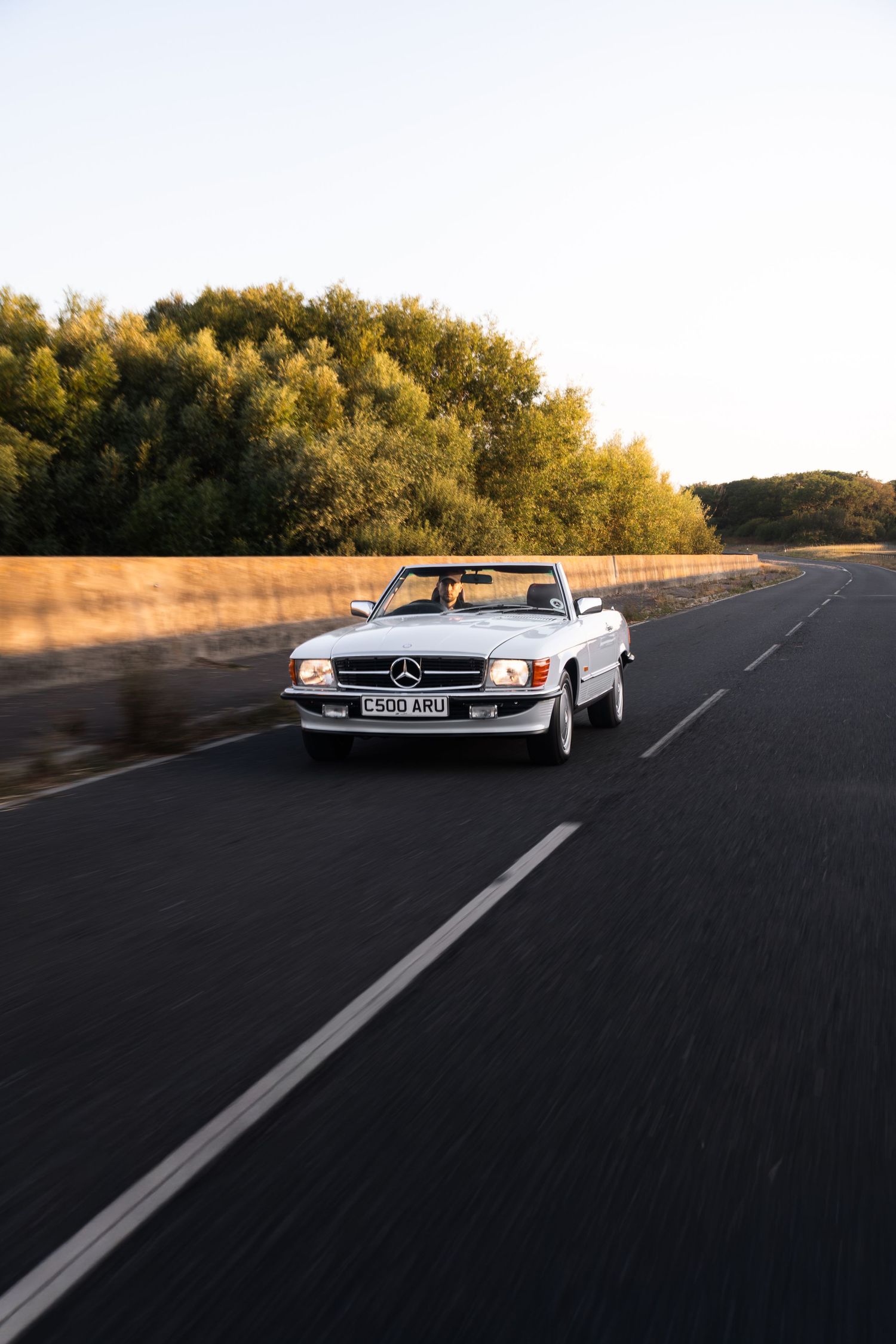White Mercedes-Benz convertible driving on open road at golden hour, surrounded by lush trees and golden fields.