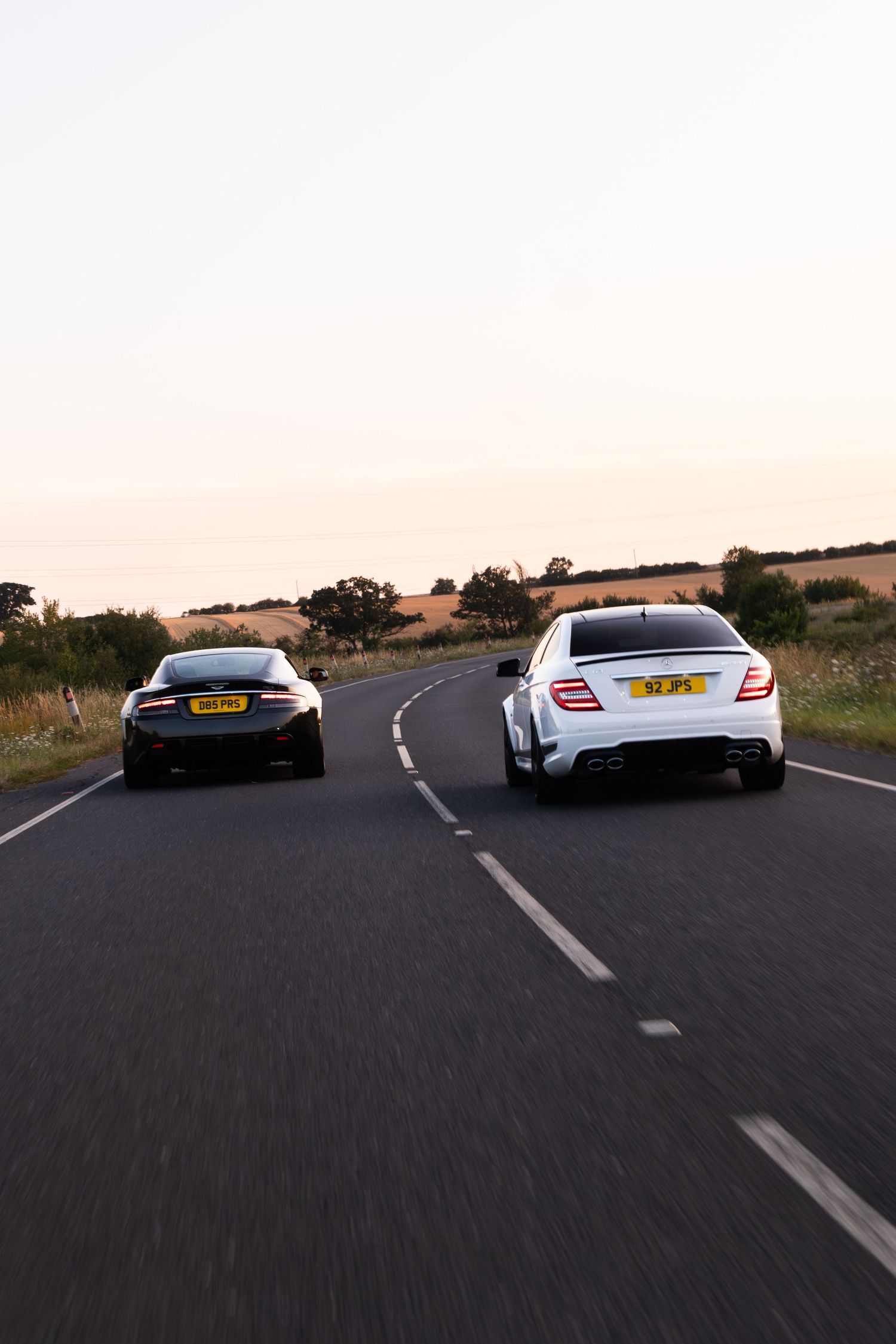 Two luxury sports cars racing on a rural UK road at dusk, viewed from behind on a winding country highway.
