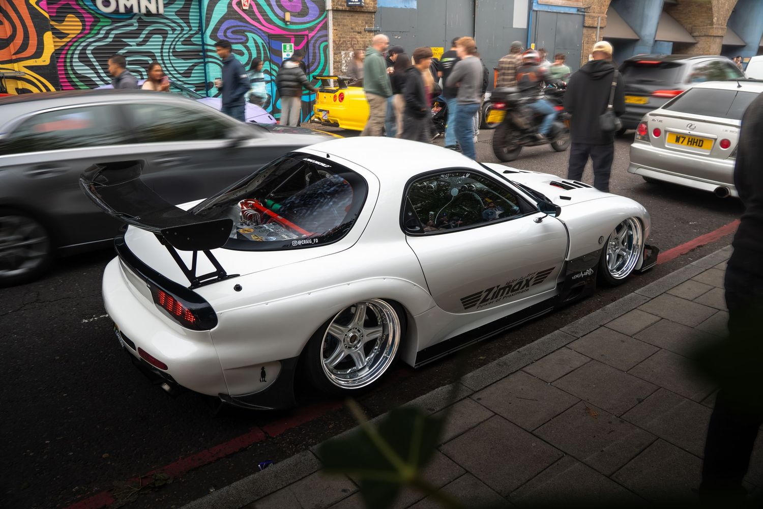 White Mazda RX-7 FD with large rear wing and wide-body kit at a car meet.