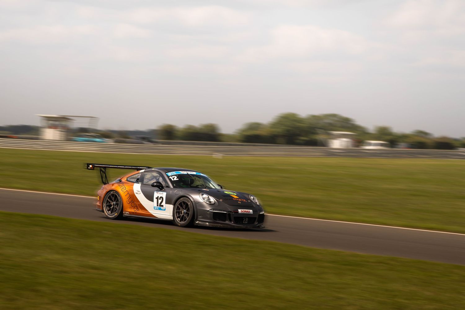 Orange and white Porsche GT3 number 12 racing on a circuit at dusk.