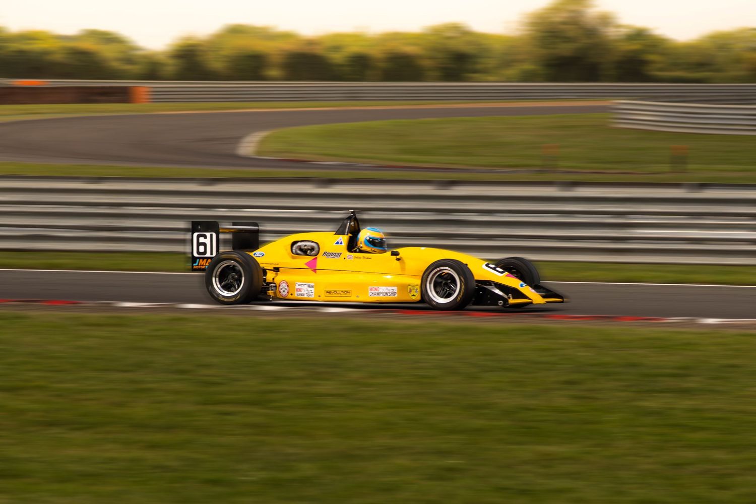 Yellow formula racing car number 61 speeding around a sunlit motorsport circuit.