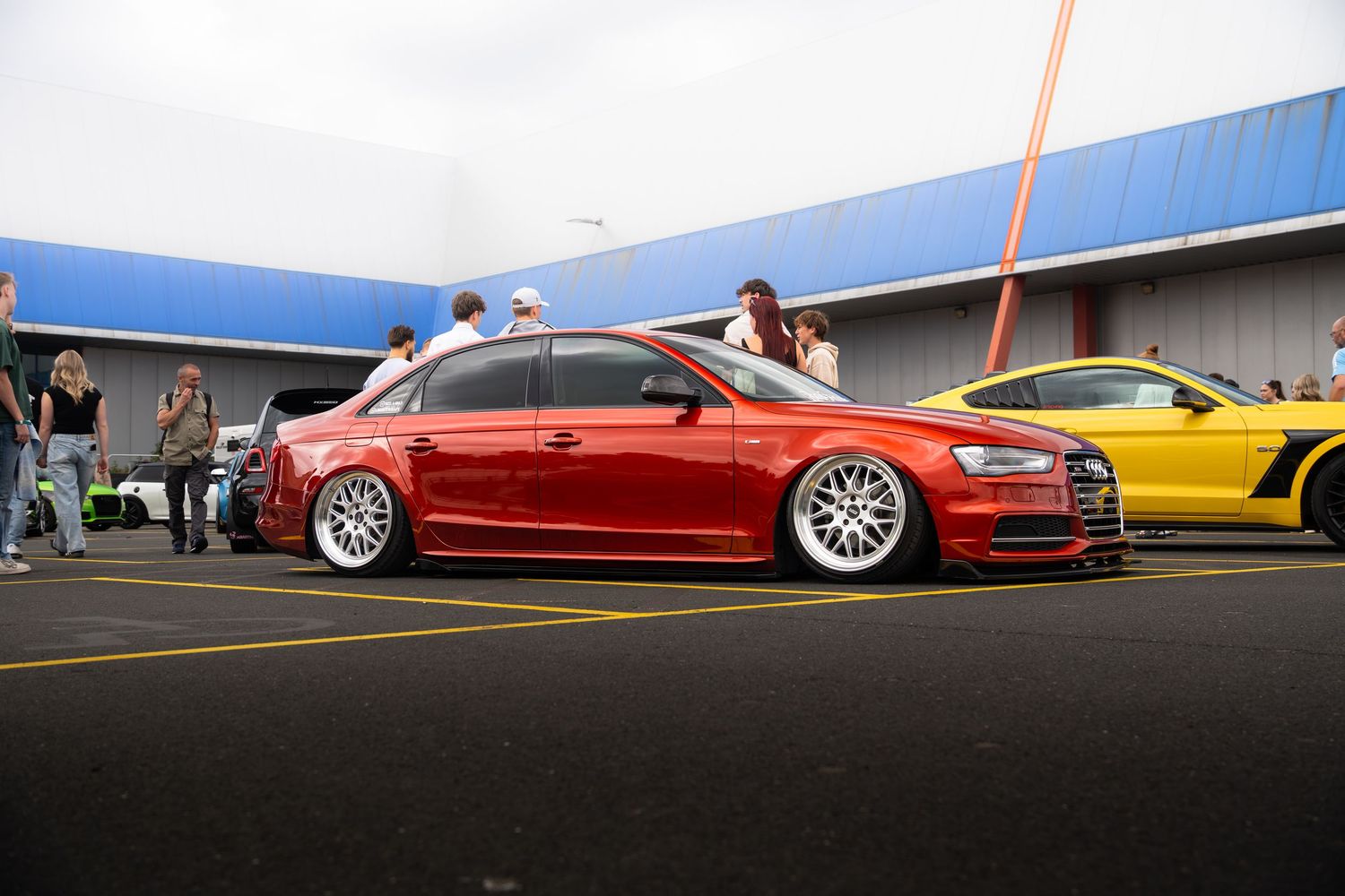Slammed red Audi S4 sedan on white deep-dish wheels parked beside a yellow sports car at a meet.