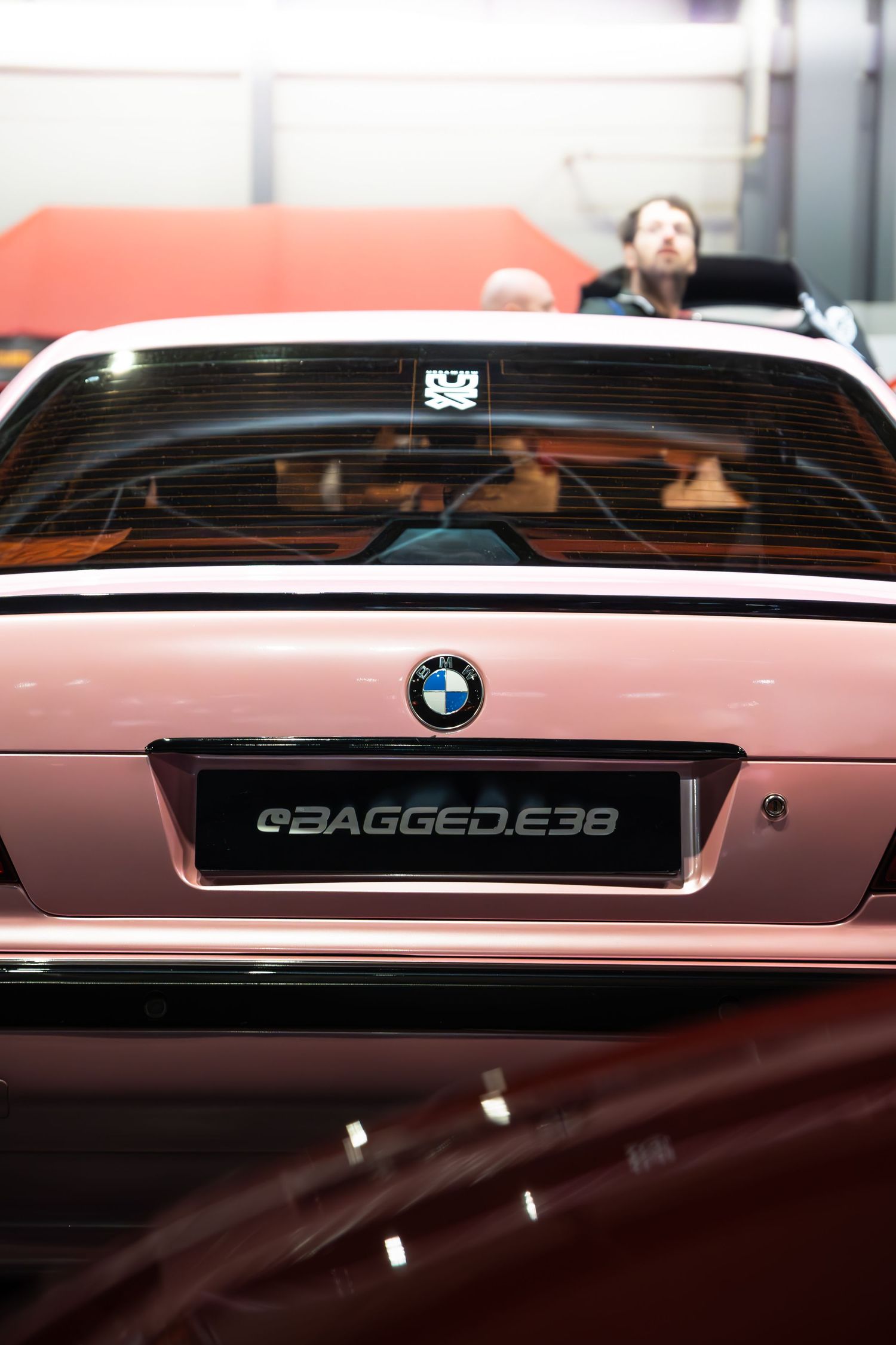 Rear view of a pink BMW E38 7 Series with BAGGED.E38 license plate at car show.