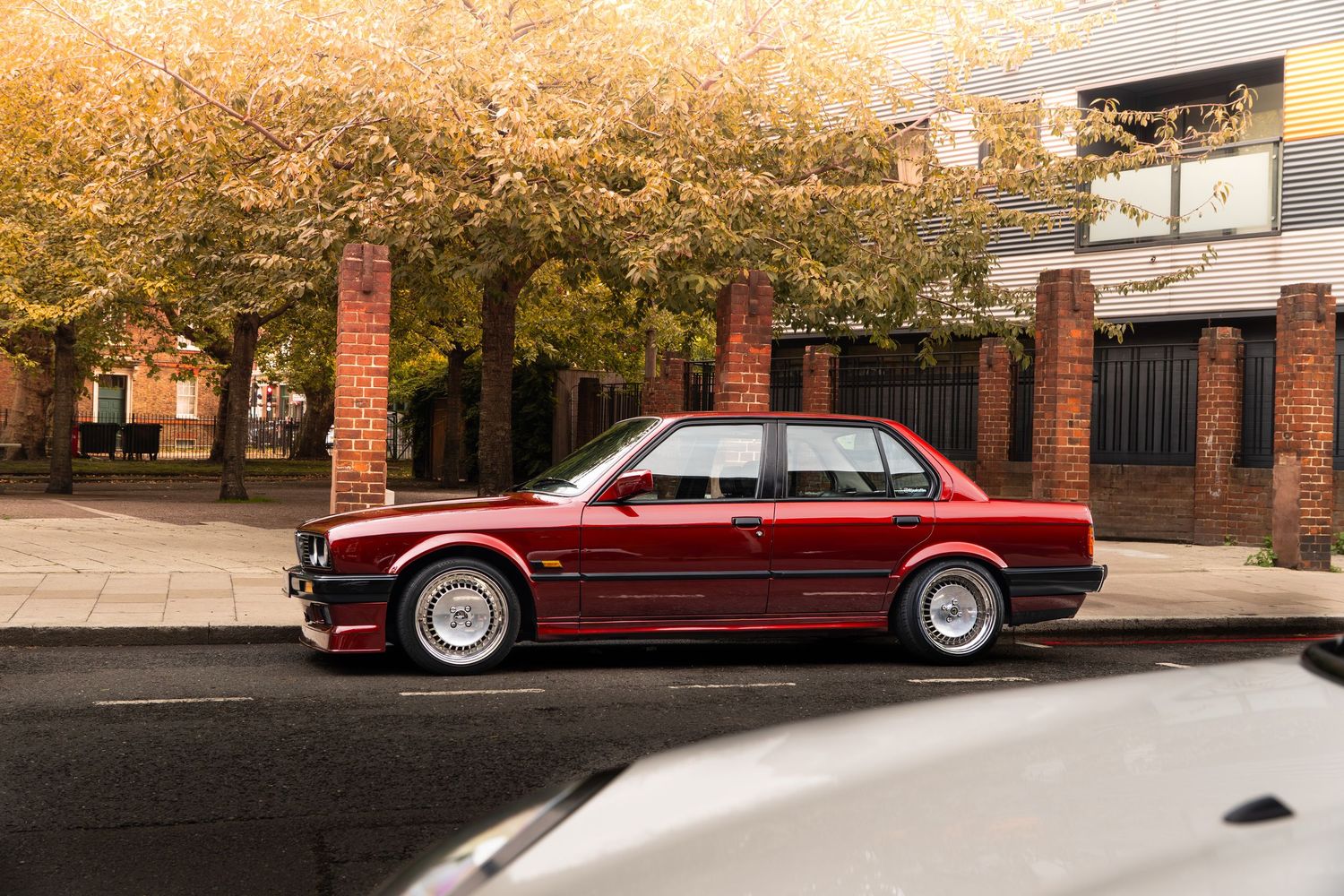 A dark red BMW E30 sedan parked on a sunlit tree-lined street in autumn.