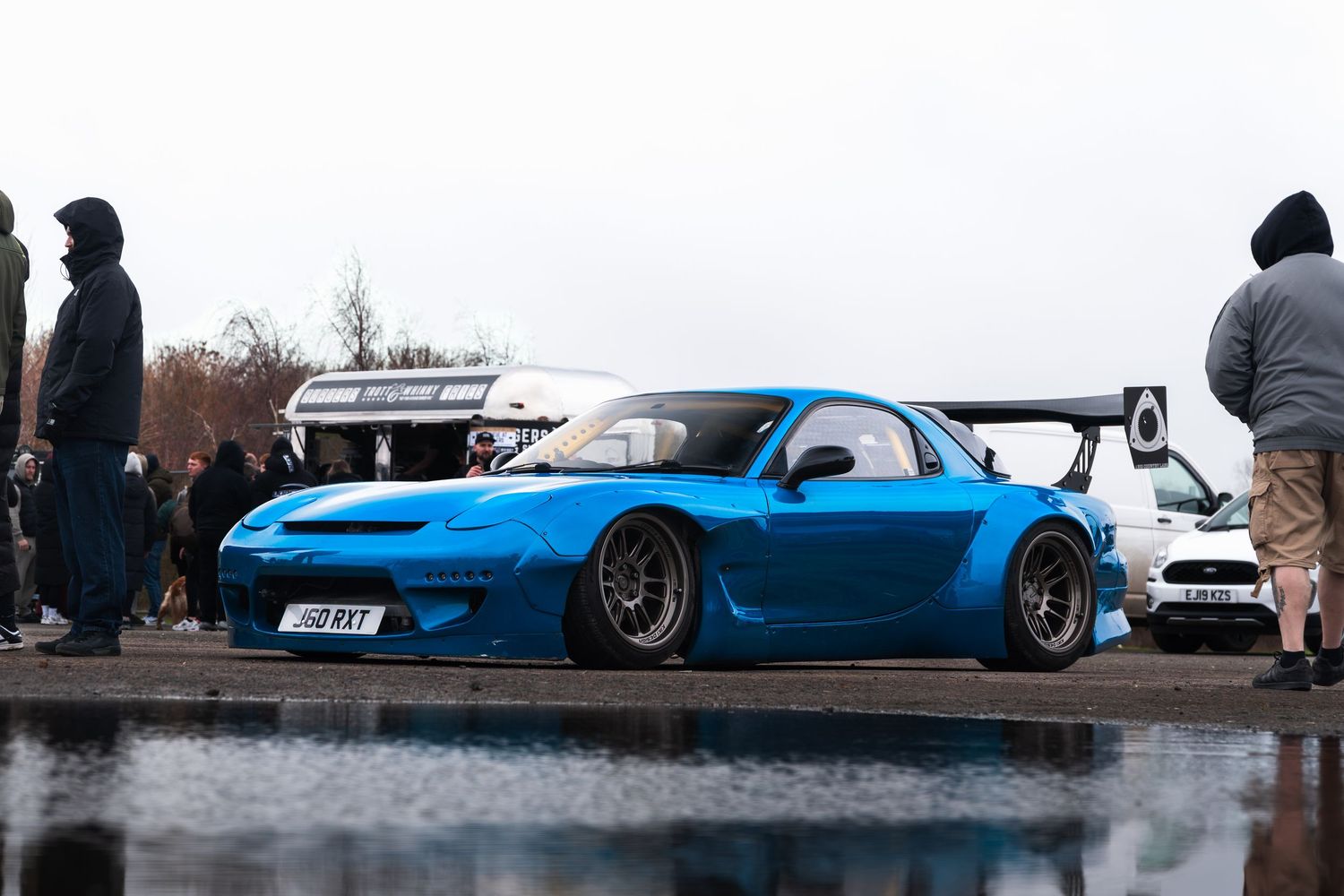 Modified blue Mazda RX-7 with wide body kit and large rear wing at a wet car meet.