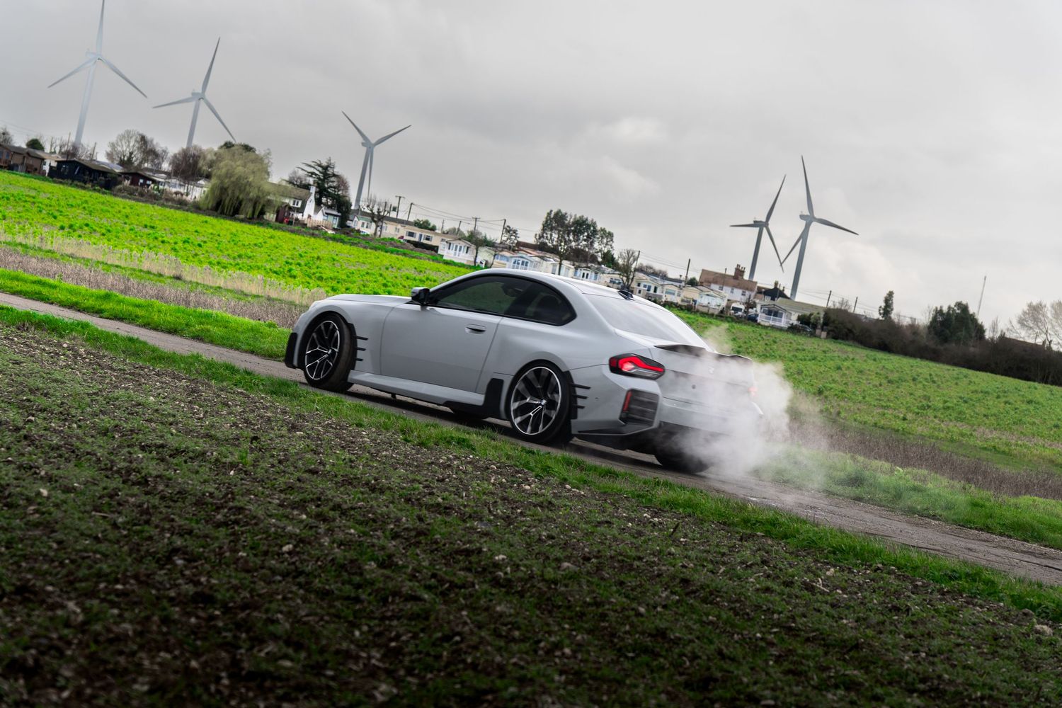 Silver BMW M2 coupe drifting with tire smoke on a rural road surrounded by green fields and wind turbines.
