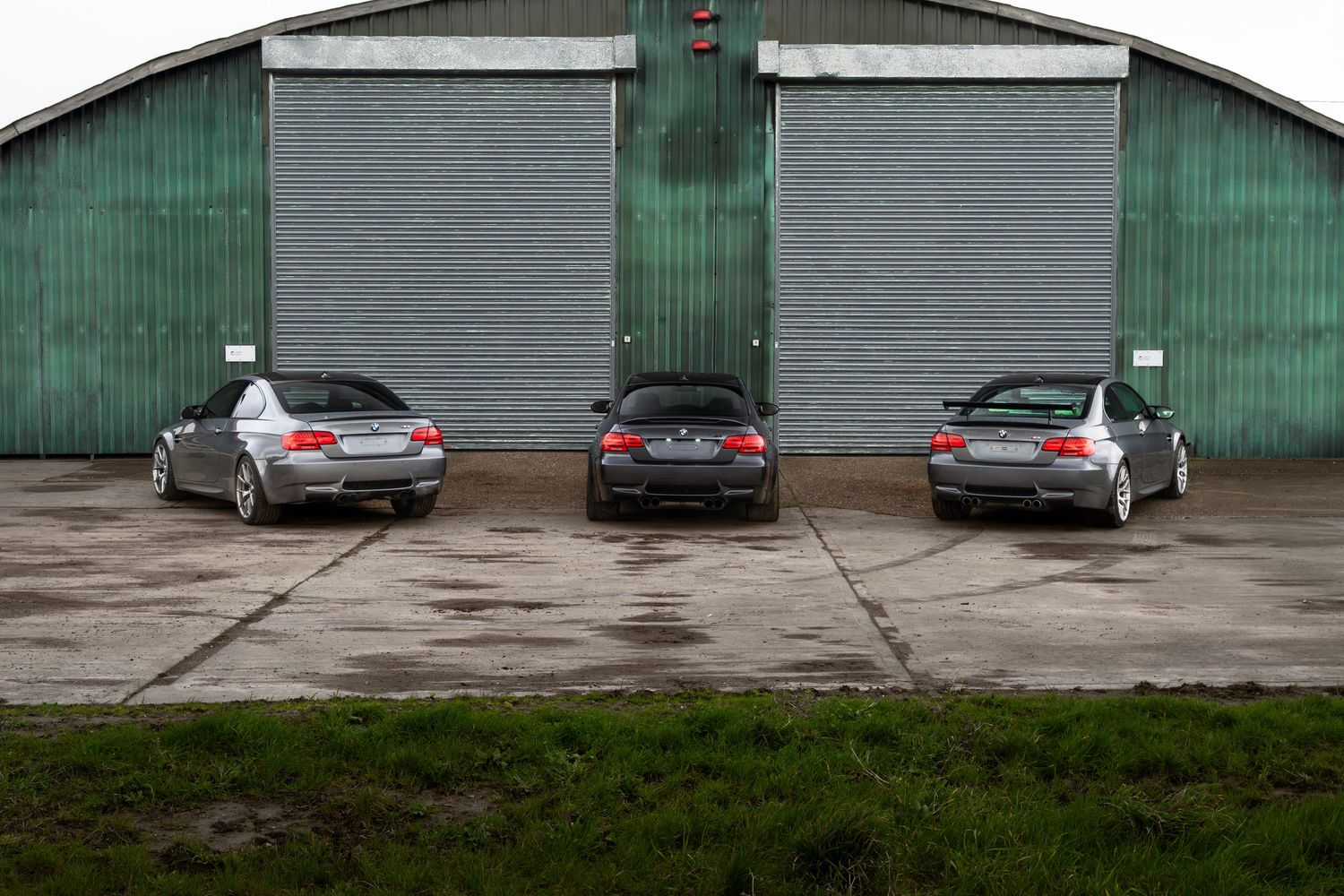 Close rear view of three BMW M3 coupes parked symmetrically in front of green garage doors.