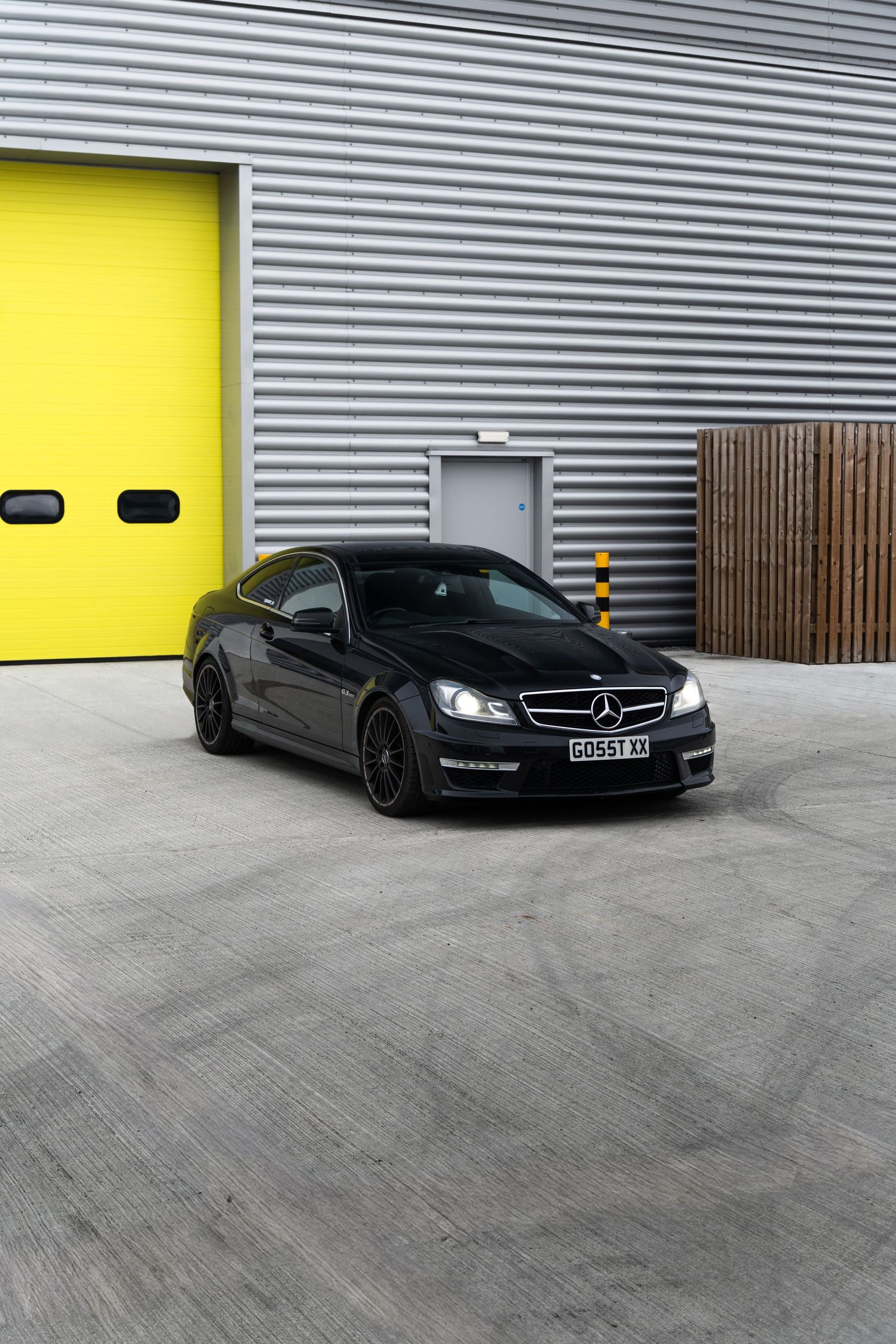 Black Mercedes-AMG C63 coupe parked in front of yellow industrial garage doors on concrete.