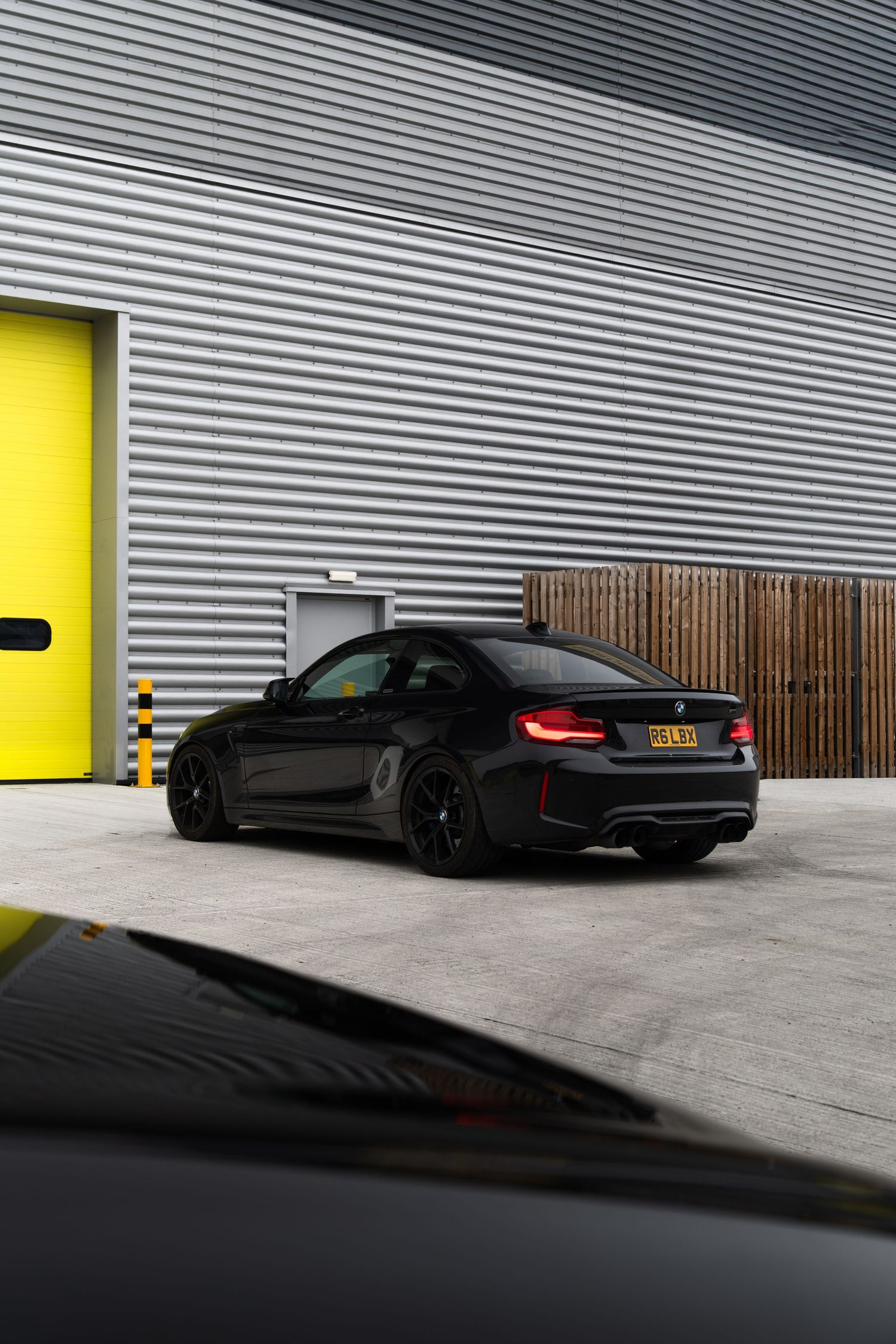Black BMW M2 coupe parked beside corrugated metal wall with yellow garage door accent.