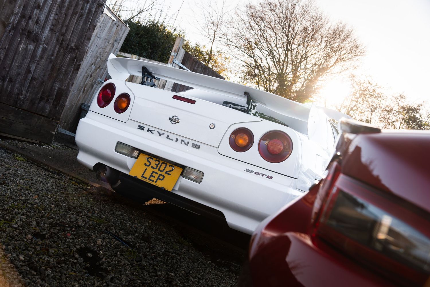 Rear view of a white Nissan Skyline R34 GTR with large spoiler and UK number plate in golden sunlight.