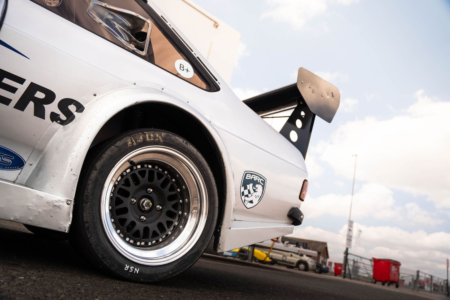 Low angle view of a white Ford RS200 rally car rear wheel and wing at a motorsport circuit.