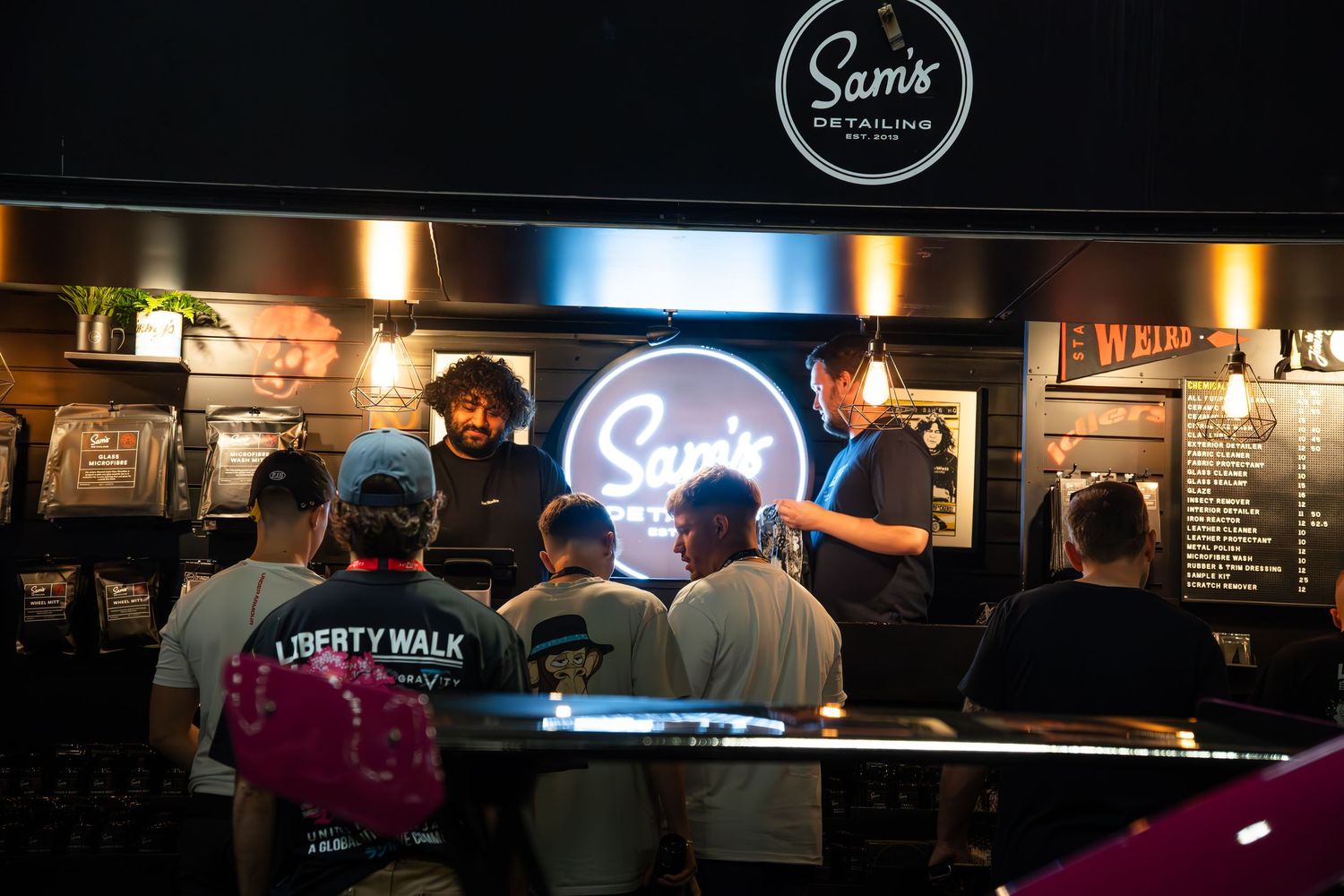 Crowd of people gathered at Sam's Detailing branded booth with neon sign at a car show event.