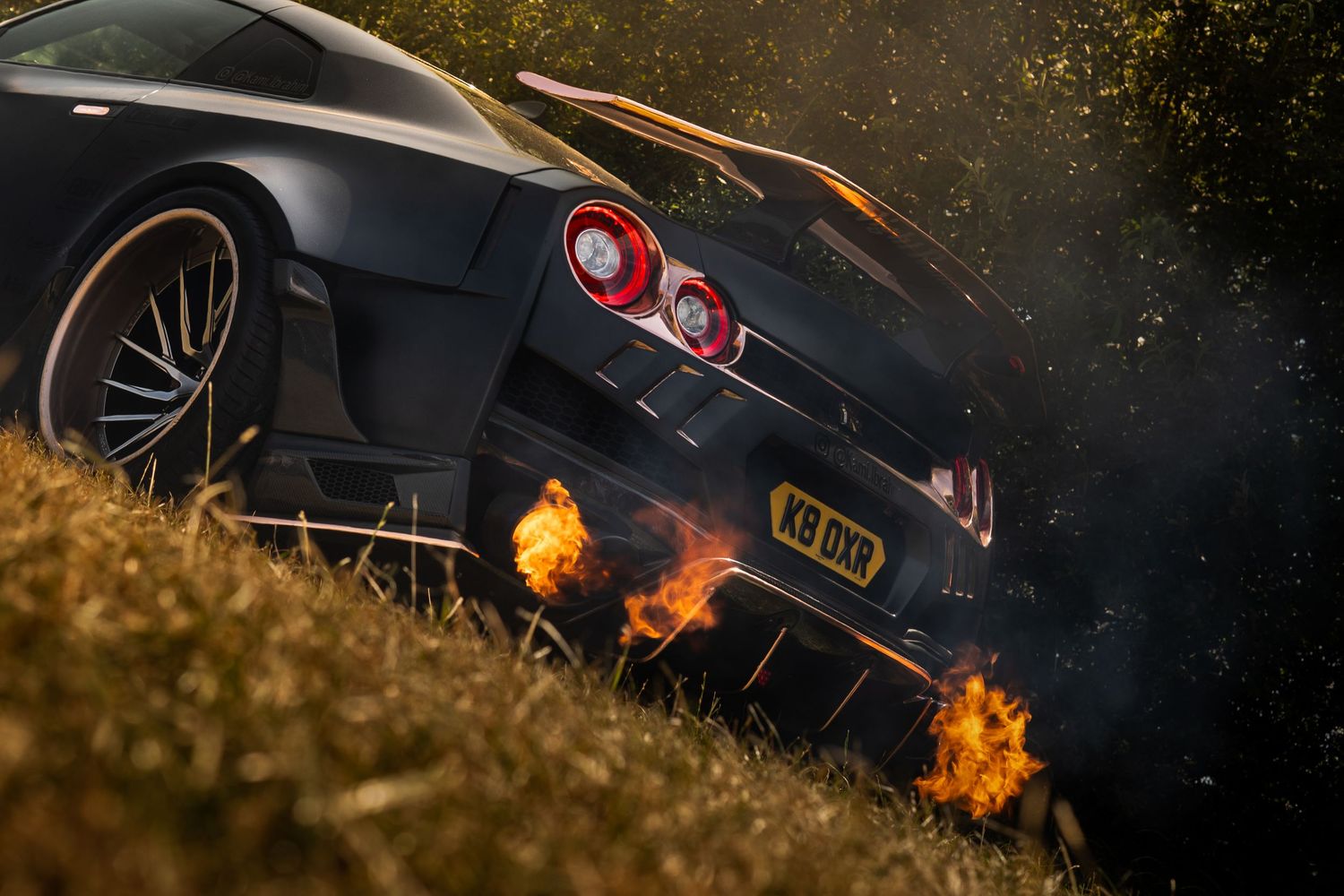 Matte black supercar shooting flames from exhaust pipes at dusk near dry grass and bushes.