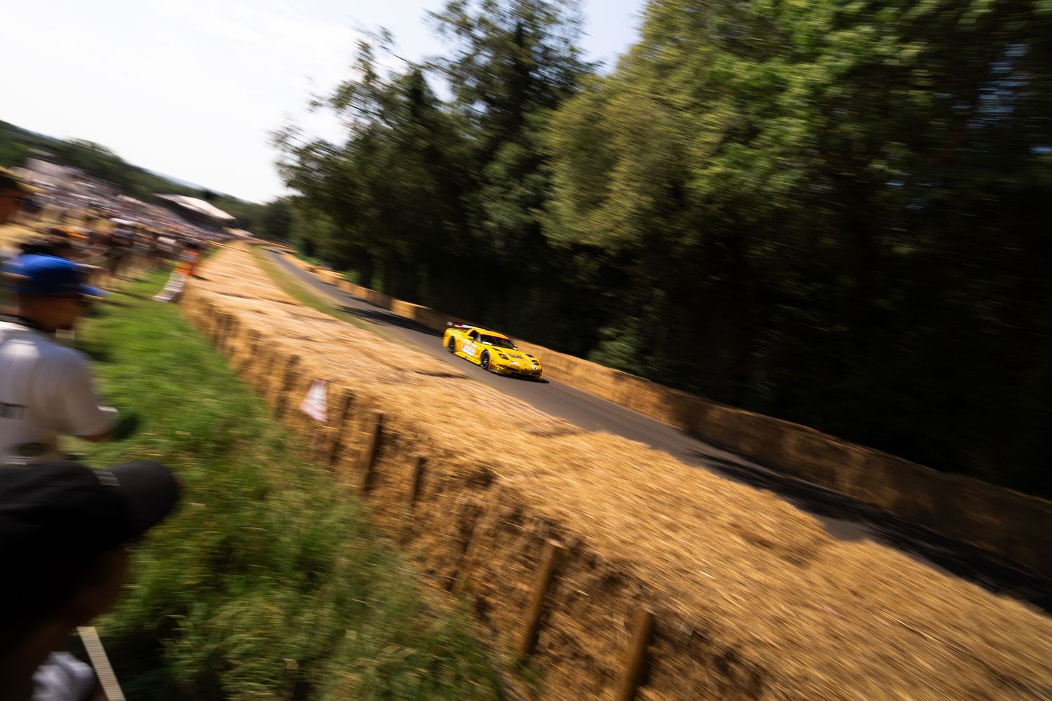 Yellow sports car racing up a hillclimb track lined with hay bales and trees at Goodwood Festival of Speed.